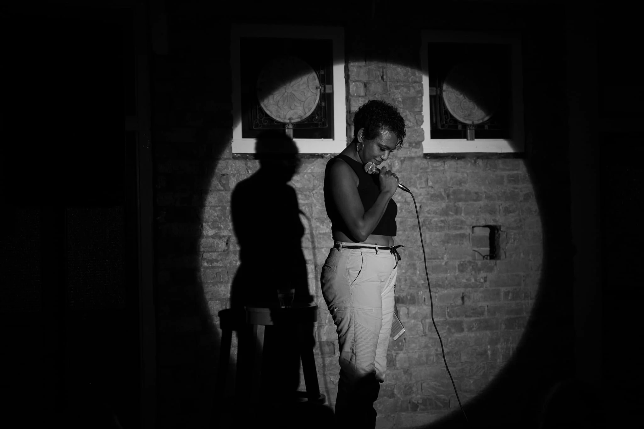 A woman stands on stage holding a microphone, looking down, with a brick wall background. Shadows are cast on the wall.