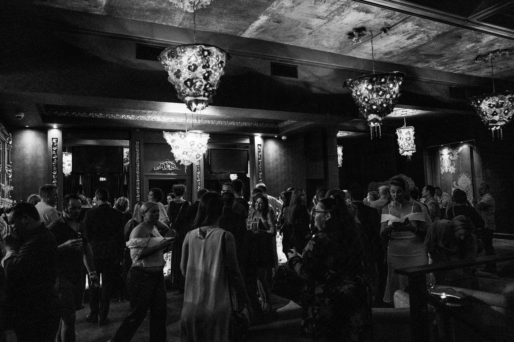 A crowded upscale indoor event with many people socializing, illuminated by ornate chandeliers hanging from a textured ceiling in a richly decorated room.