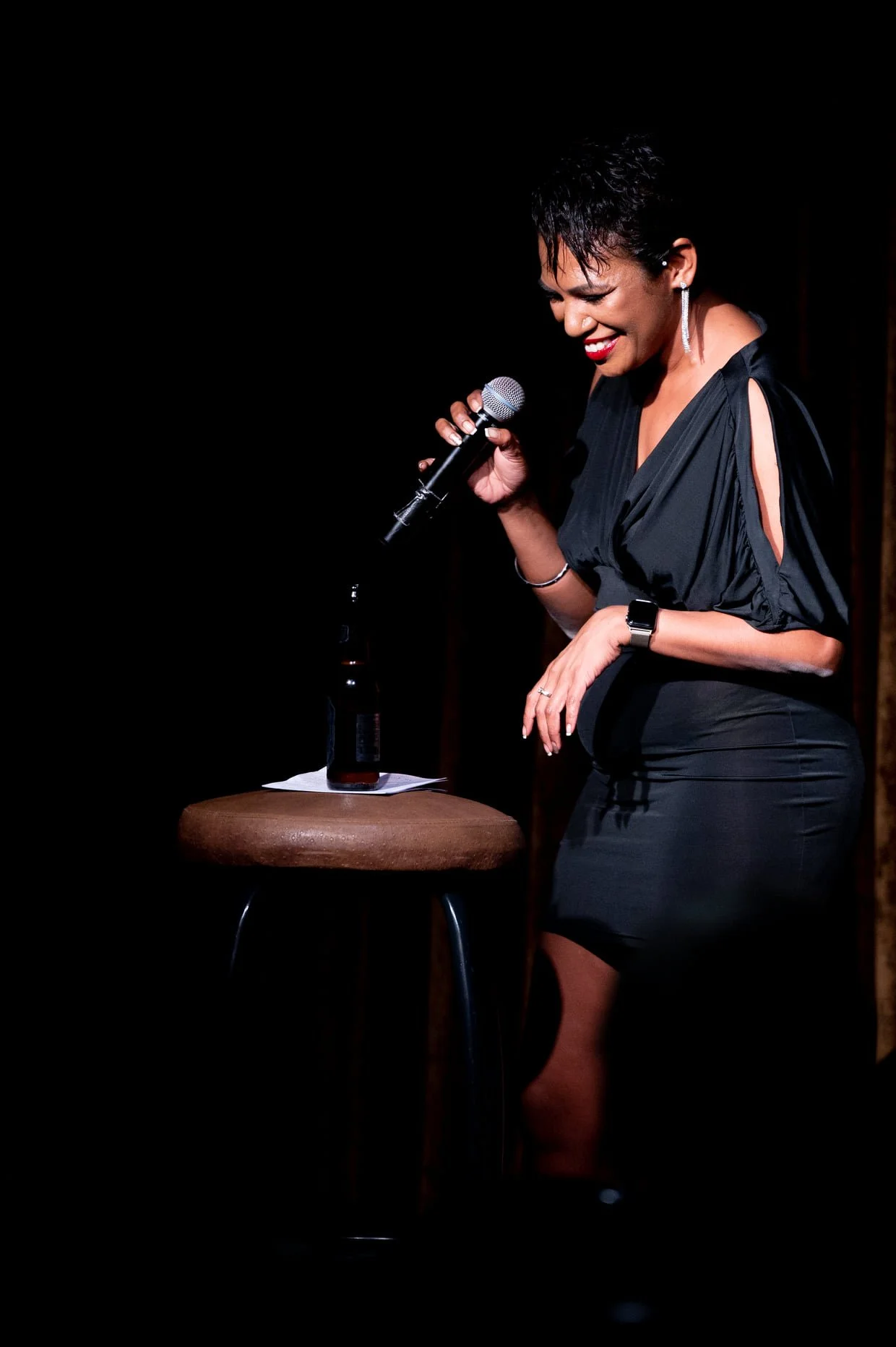 A woman with short, dark hair and earrings, wearing a black dress, is smiling and holding a microphone. She is standing near a stool with a beer bottle on it, on a dark stage or performance area.