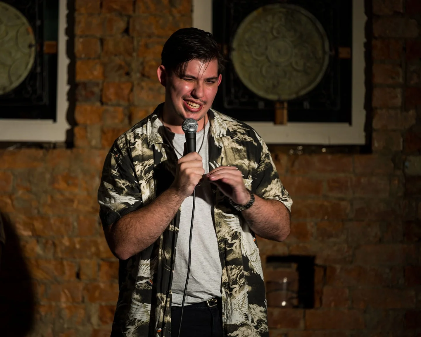 A young man performing stand-up comedy on stage, holding a microphone, wearing a patterned short-sleeve shirt and a watch, with a brick wall and framed pictures in the background.