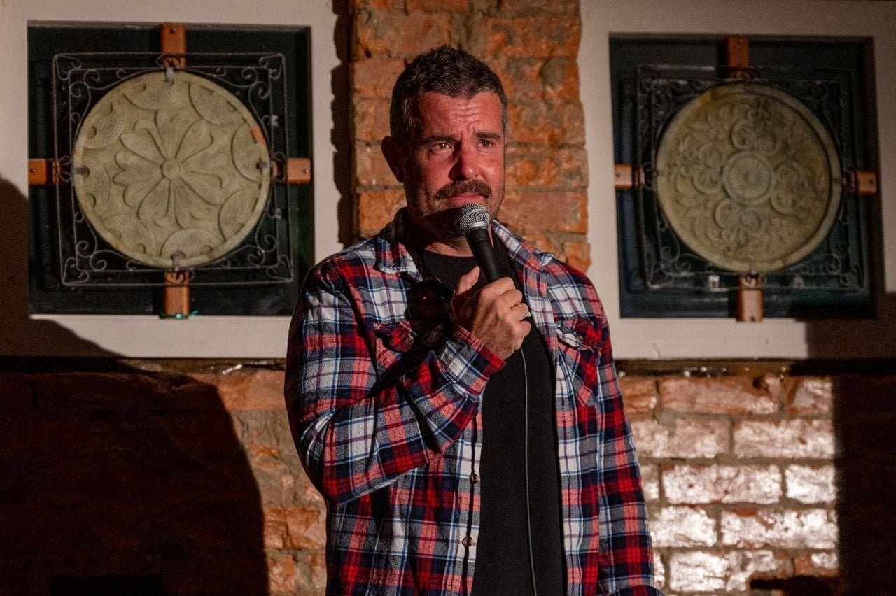 A man with a beard and short hair holding a microphone and performing stand-up comedy in a dimly lit venue with bricks and decorative wall art in the background.