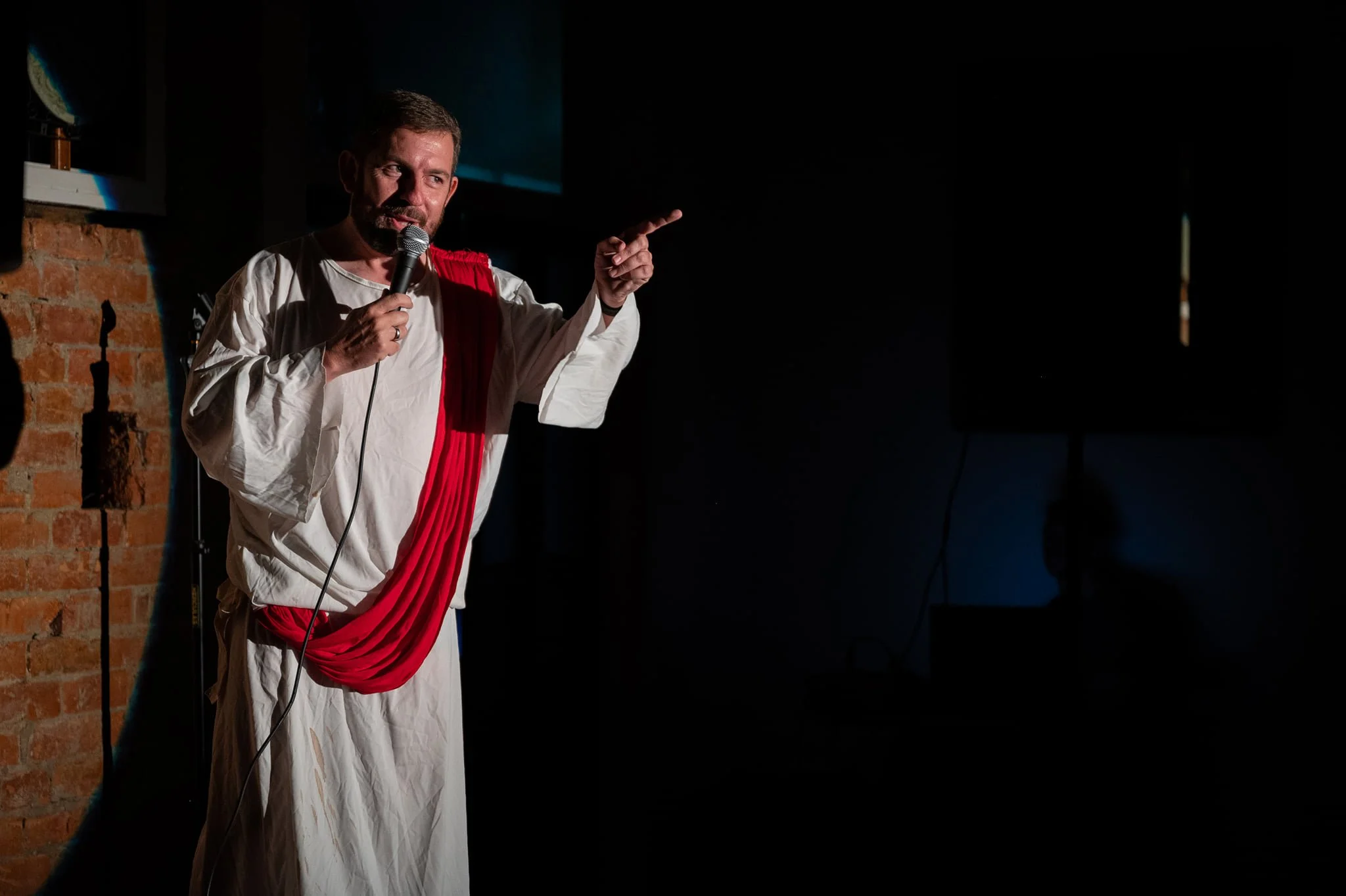 Man dressed as a Roman emperor or senator, holding a microphone and pointing, standing in front of a brick wall and a dark background.