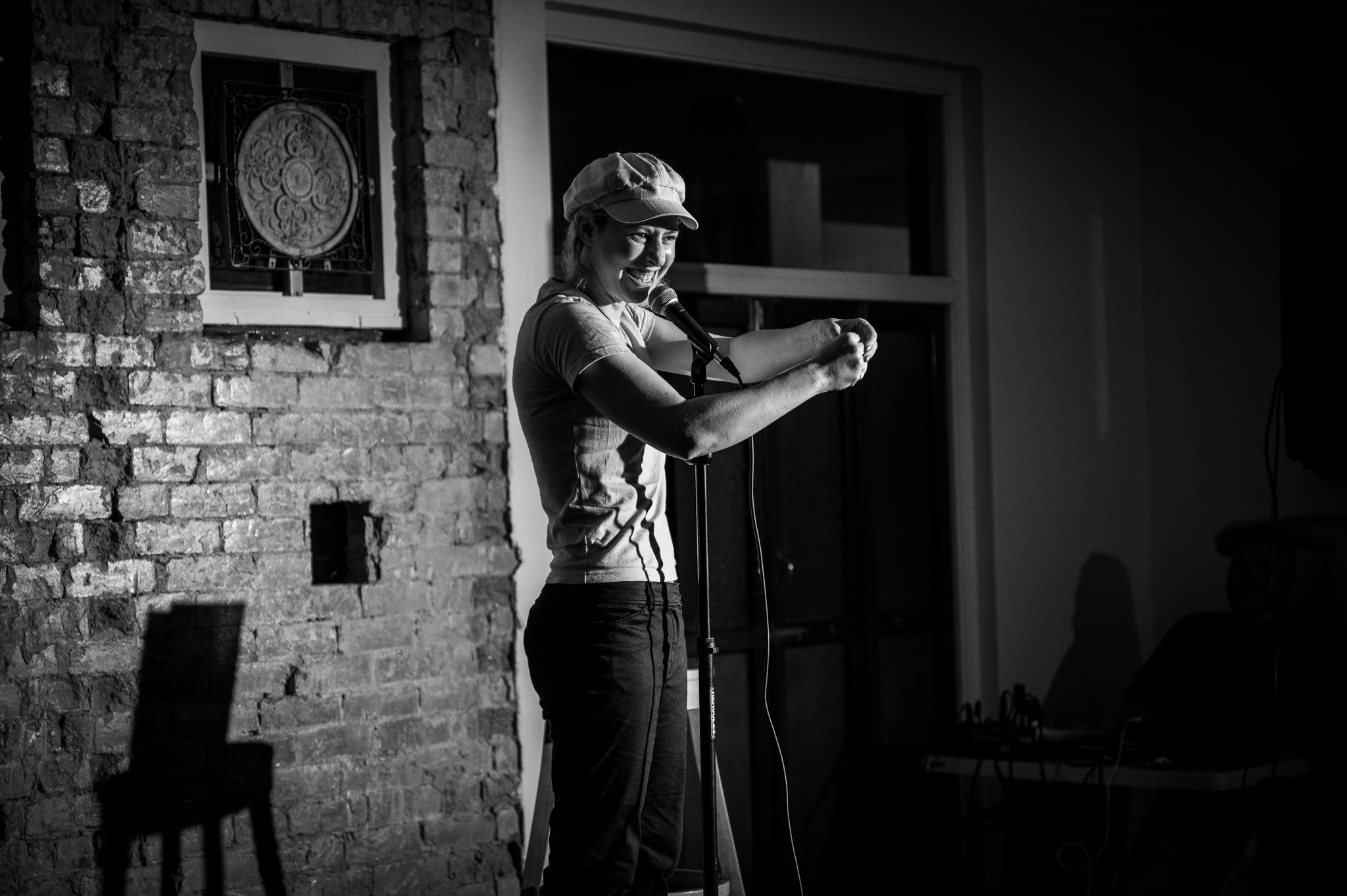 A woman performing stand-up comedy on stage, smiling and laughing, with a brick wall and a window behind her, holding a microphone.