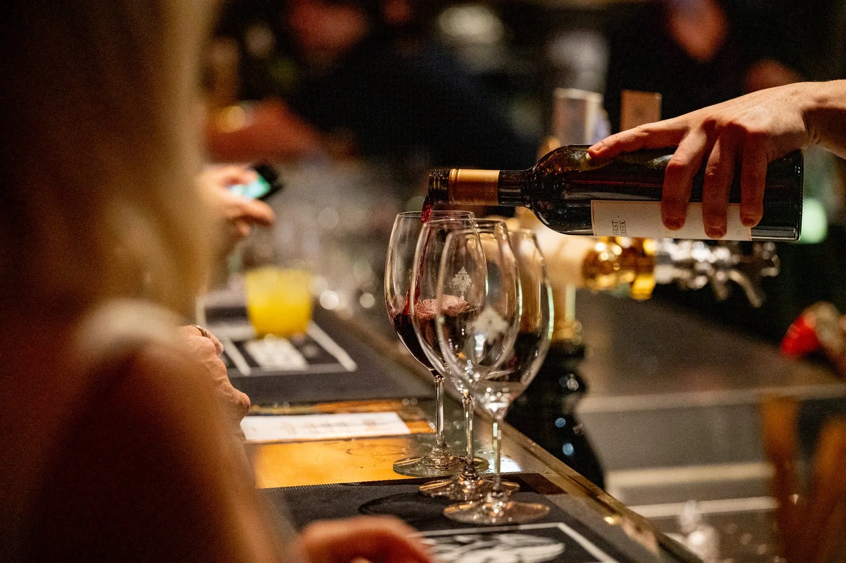 A person is pouring red wine from a bottle into a wine glass on a bar counter, with another wine glass already filled nearby. The background is blurred, showing a dimly lit bar setting.