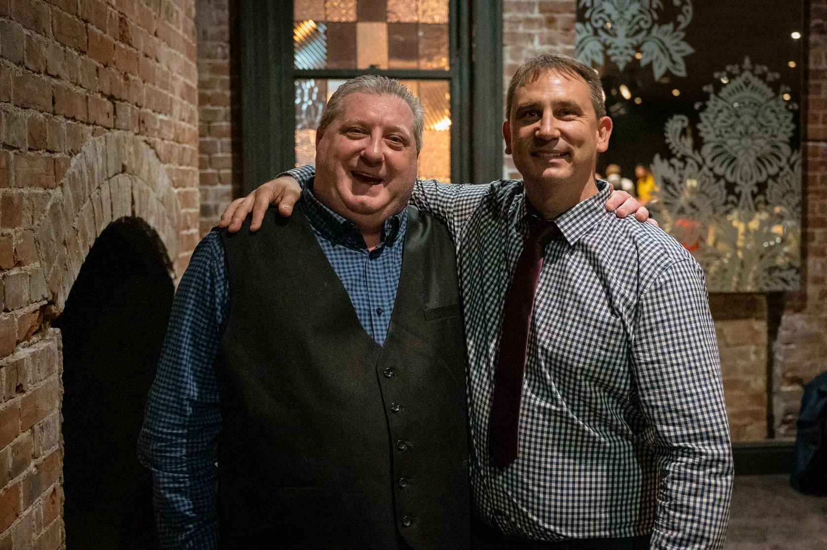 Two men smiling and standing together with their arms around each other's shoulders in a dimly lit restaurant or bar, with brick walls and decorative patterns in the background.