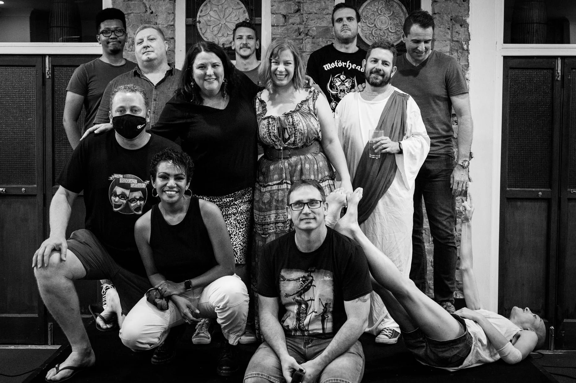 Group of ten people, some smiling, some serious, posing for a black-and-white photo indoors in front of a brick wall with decorative metal accents. Two people are sitting on the floor, four are kneeling or squatting, and four are standing. One person