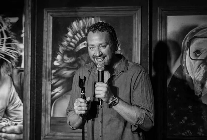 Man smiling while holding a microphone, standing in front of framed artwork on a wall.