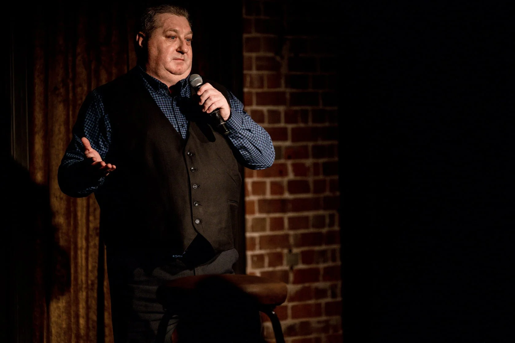 A man standing on stage holding a microphone, wearing a dark vest and checkered shirt, with a brick wall backdrop.