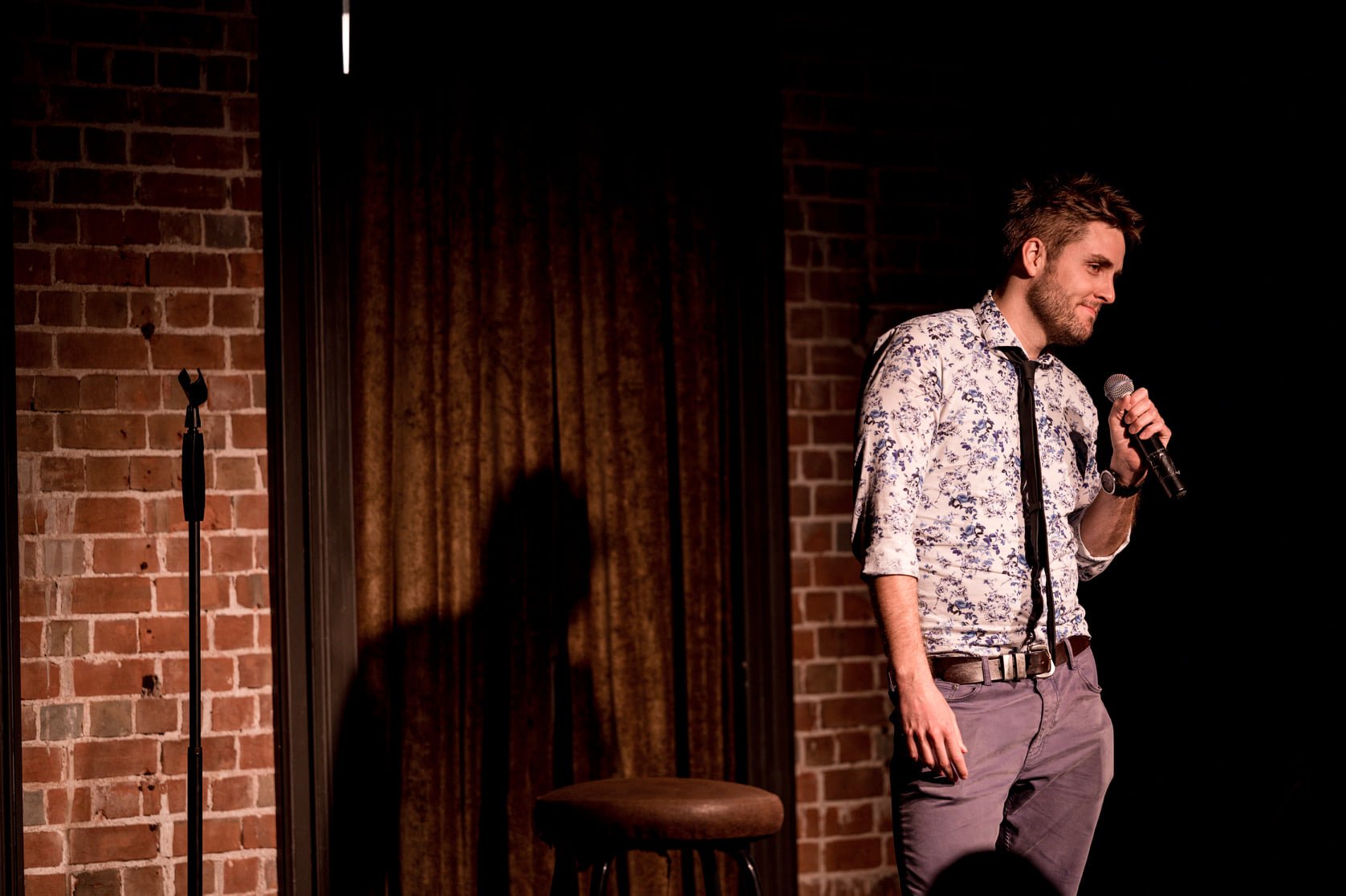 A man in a floral shirt holding a microphone on a stand-up comedy stage with brick walls and a curtain in the background.