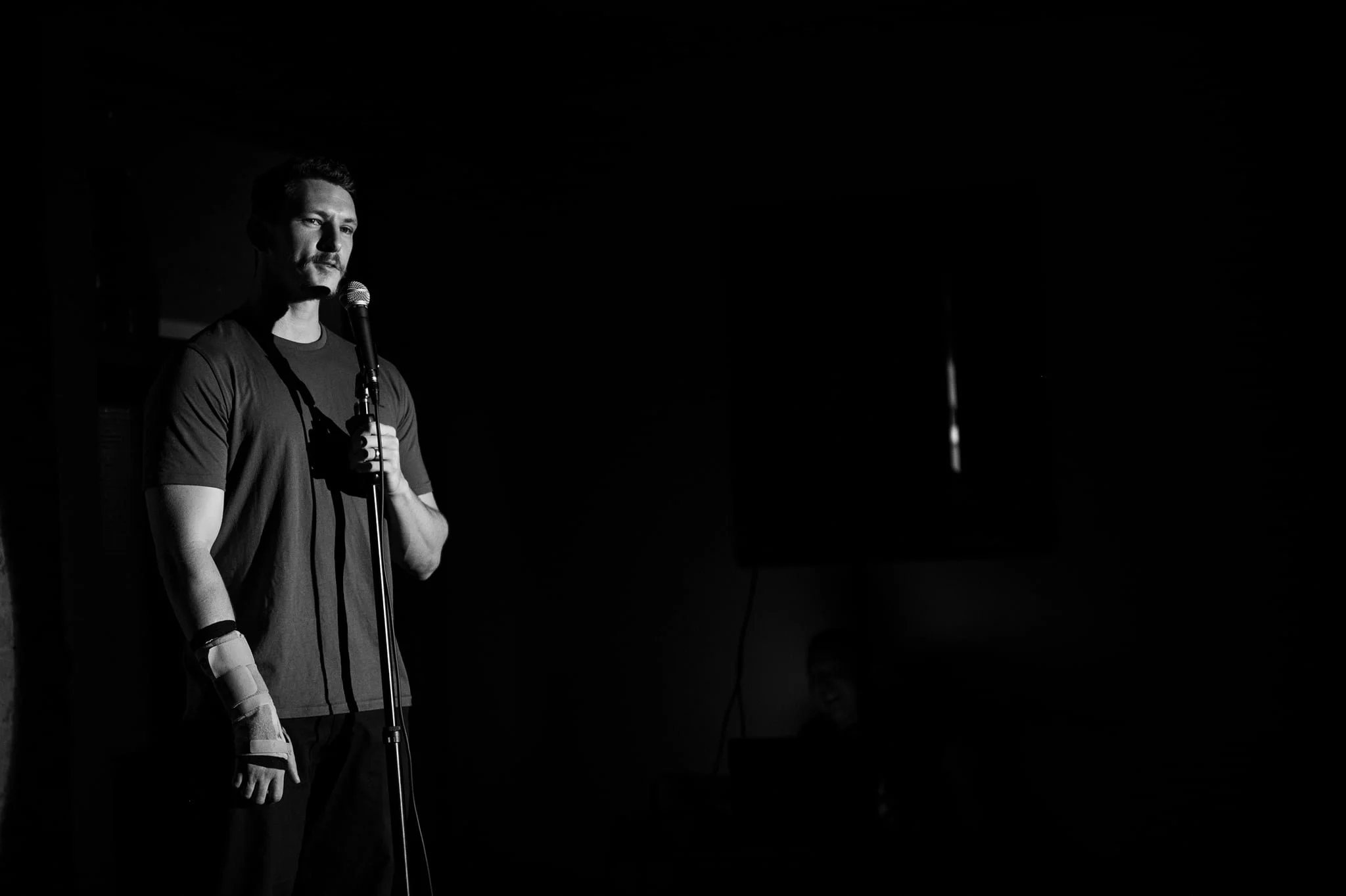 A man stands alone on stage holding a microphone in front of a dark background, with a serious expression.