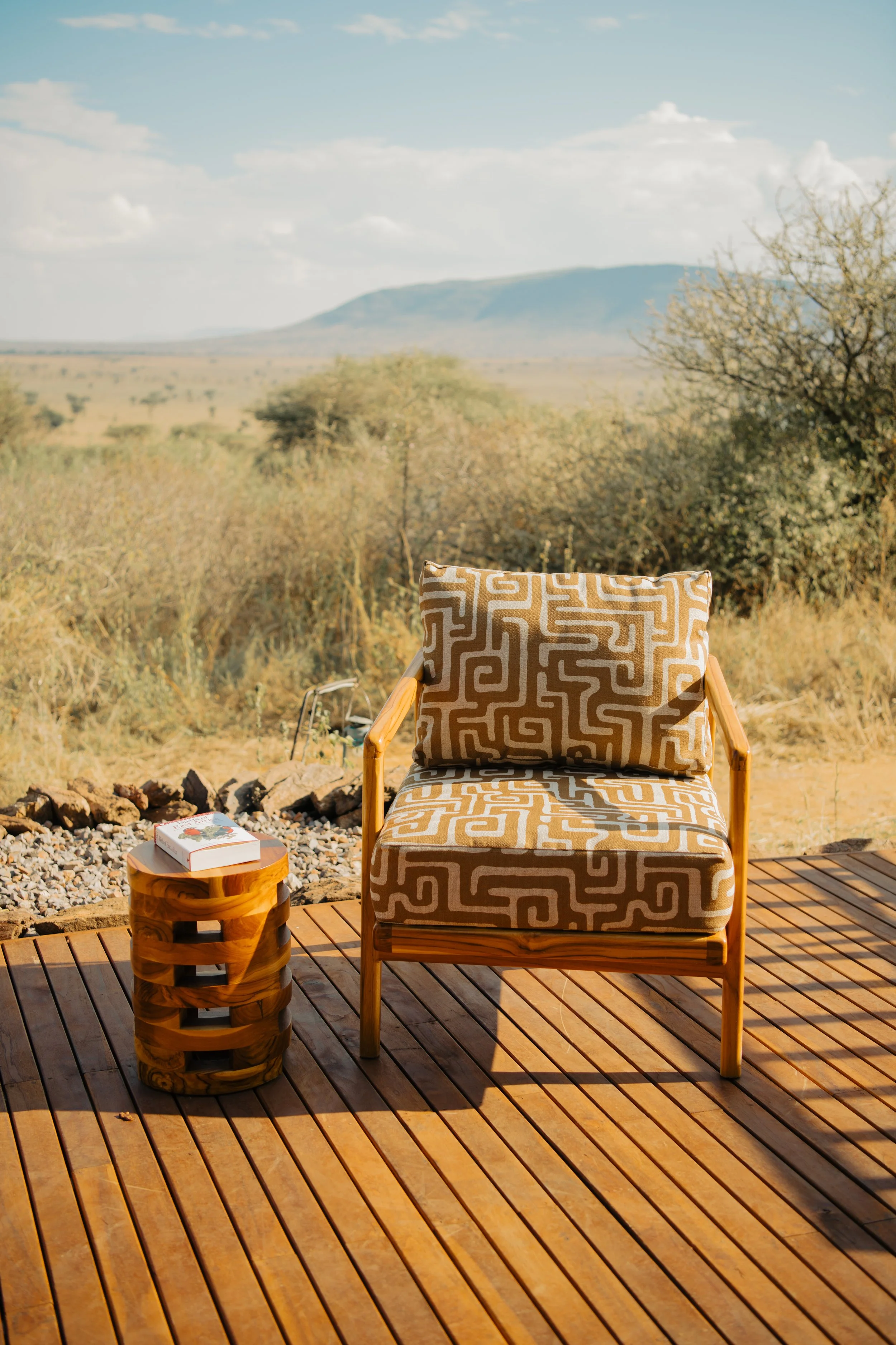 Kisasa Designs wooden chair with patterned cushions on a deck, side table with a book, overlooking a savanna landscape.