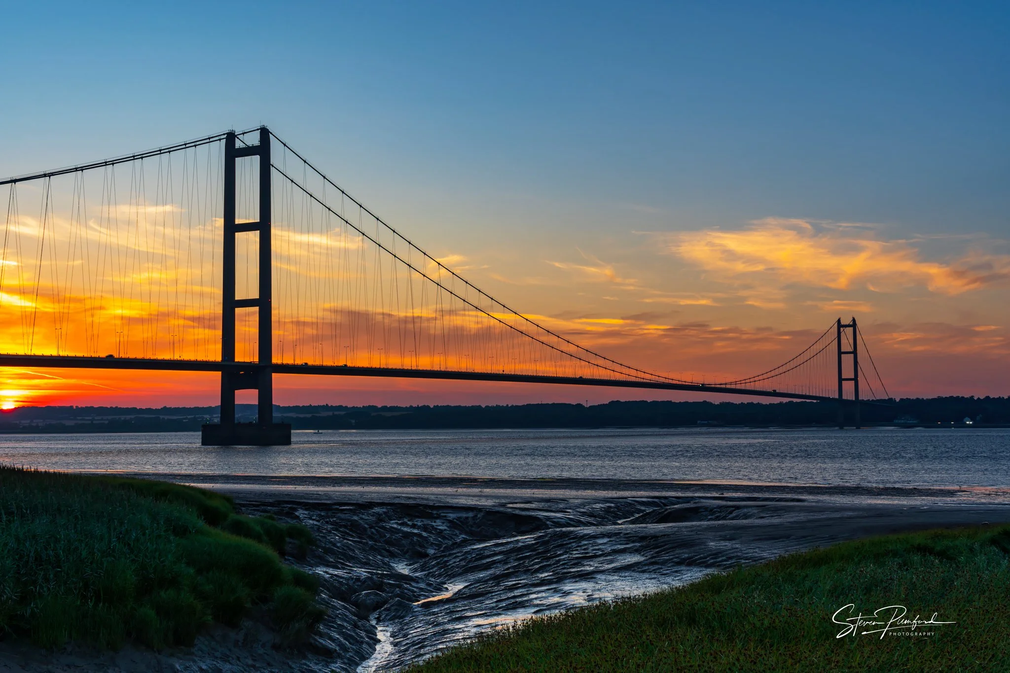 The Humber bridge spans over the Humber Estuary during sunset, with a colorful sky and clouds in the background.