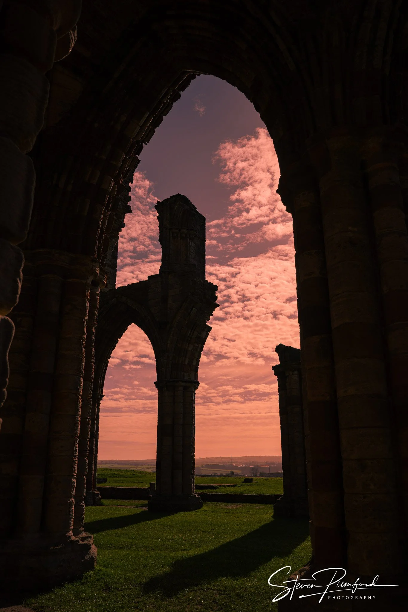 Ancient stone ruins with tall arches and columns against a pink sunset sky, with a field and distant horizon visible through the arches.