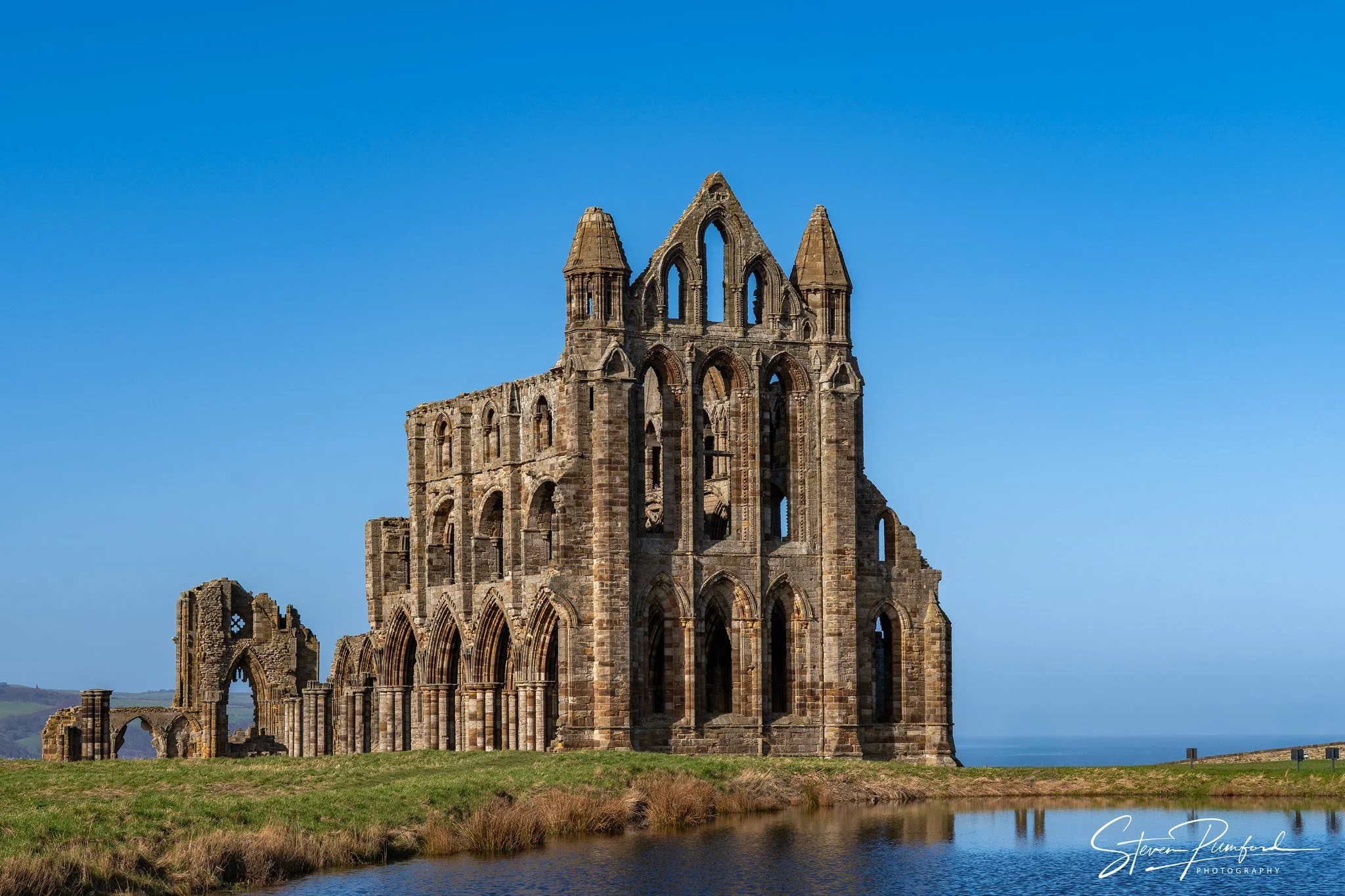 Ruined medieval stone church with tall pointed towers under a clear blue sky, surrounded by a grassy field and a small body of water in the foreground.