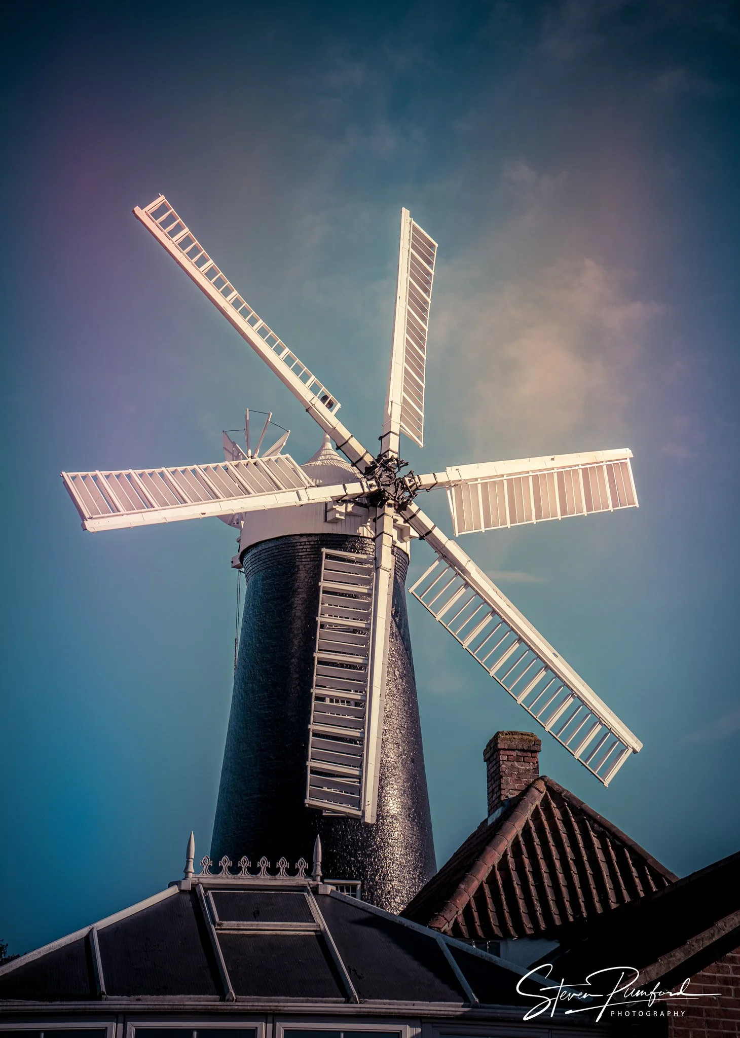 A traditional windmill with white blades and a black cylindrical body, situated next to a brick house with a tiled roof, under a blue sky.