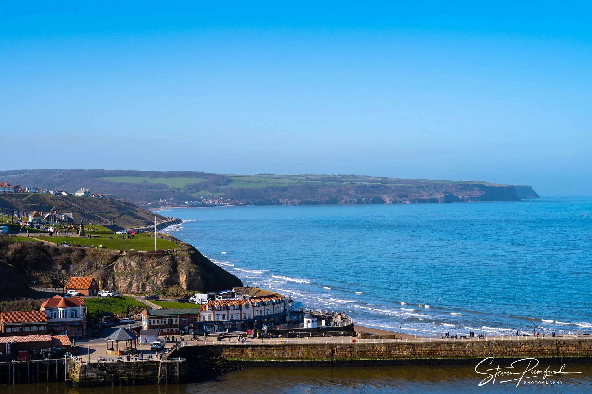 Coastal scene with a town along the shoreline, green hills in the background, and a clear blue sky.