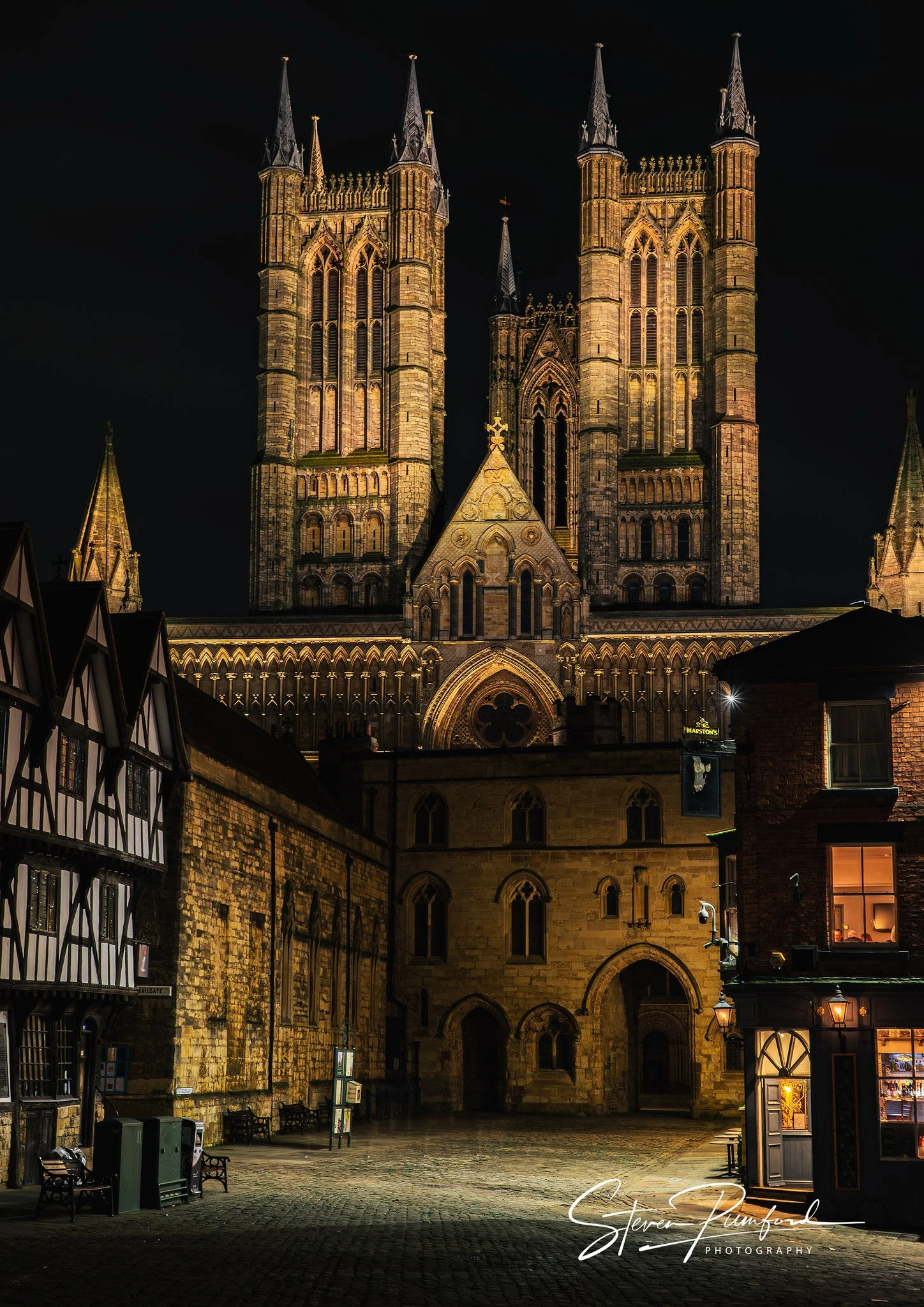 Night view of a historic Gothic-style Lincoln Cathedral illuminated, seen from a cobblestone square with benches, lampposts, and surrounding buildings. The sky is dark, highlighting the cathedral's architecture.
