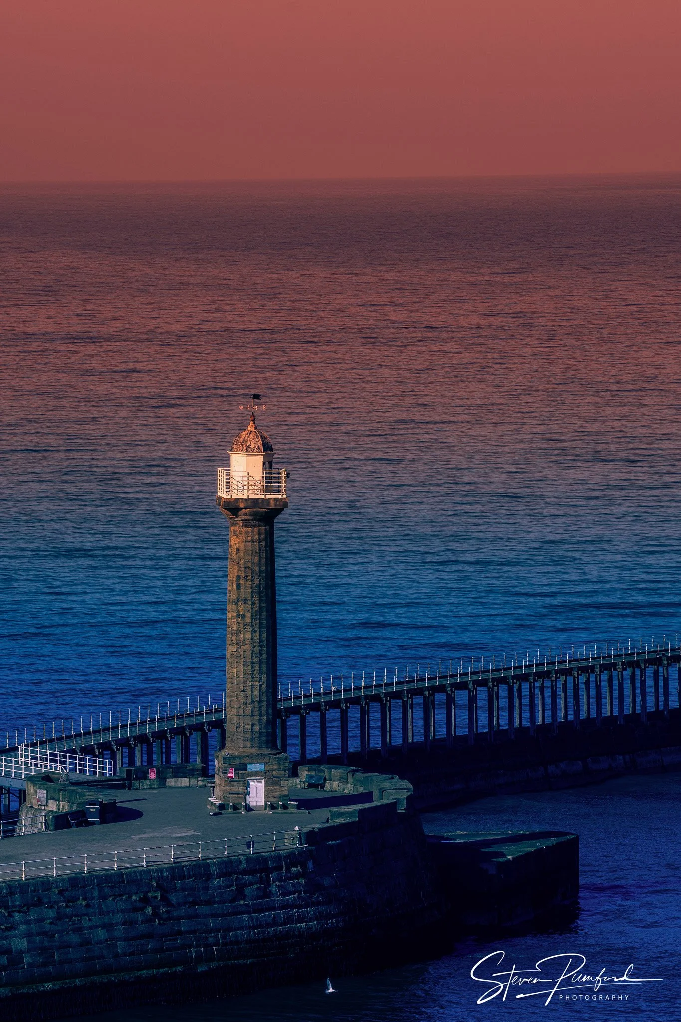 A lighthouse with a dome-shaped roof and a weather vane marking directions, situated on a stone pier extending into the ocean. The scene is during the evening with a pinkish sky and calm water, with a small sailboat visible in the water.
