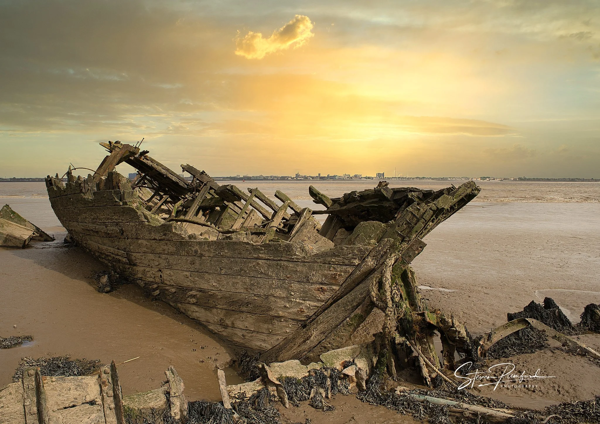 A decayed wooden boat wreck on a sandy beach during sunset with a distant city skyline in the background.