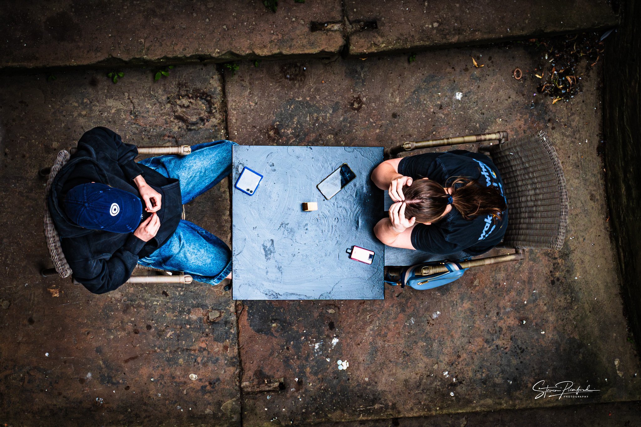Overhead view of two people sitting at a small, rectangular outdoor table on a weathered concrete surface. One person is wearing a cap and dark clothing, while the other has brown hair tied back, wearing a dark t-shirt. There are two smartphones, an 