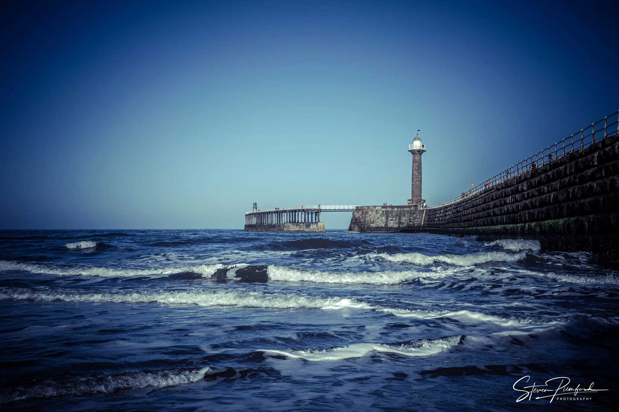 A lighthouse on a pier extending into the ocean, with waves in the foreground and a clear blue sky in the background.