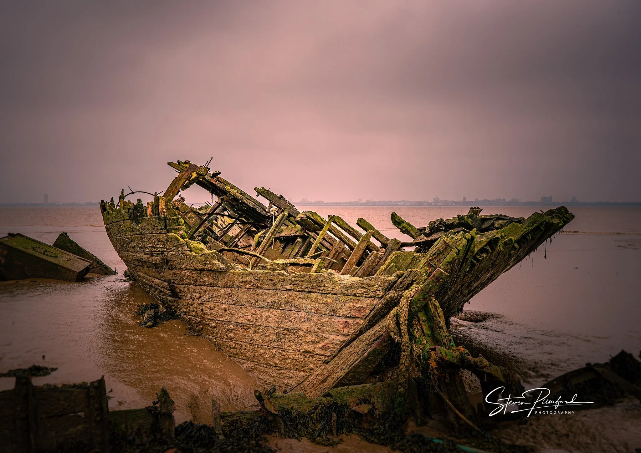 A decayed and abandoned shipwreck on a muddy shoreline with city skyline in the distance under a cloudy sky.