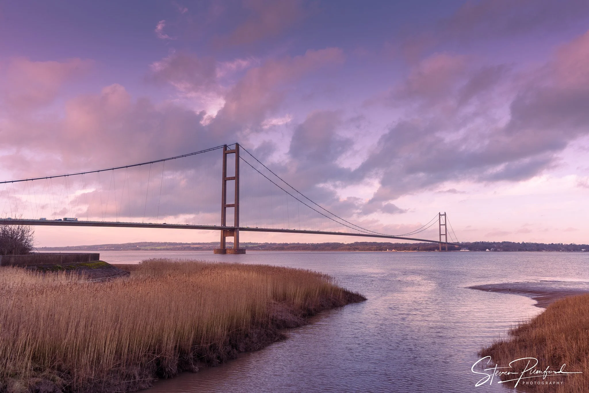 A suspension bridge over a wide river at sunset, with purple and pink clouds in the sky and grasses along the shoreline.