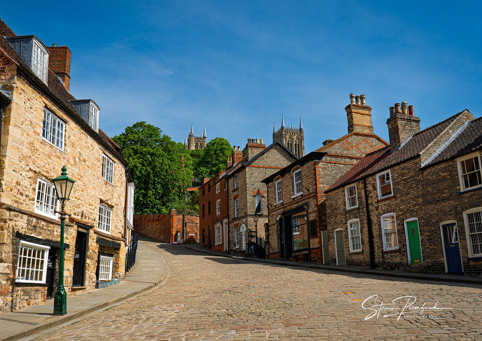 A cobblestone street in Lincoln lined with historic brick and stone houses, with a church or cathedral with tall spires in the background under a blue sky.