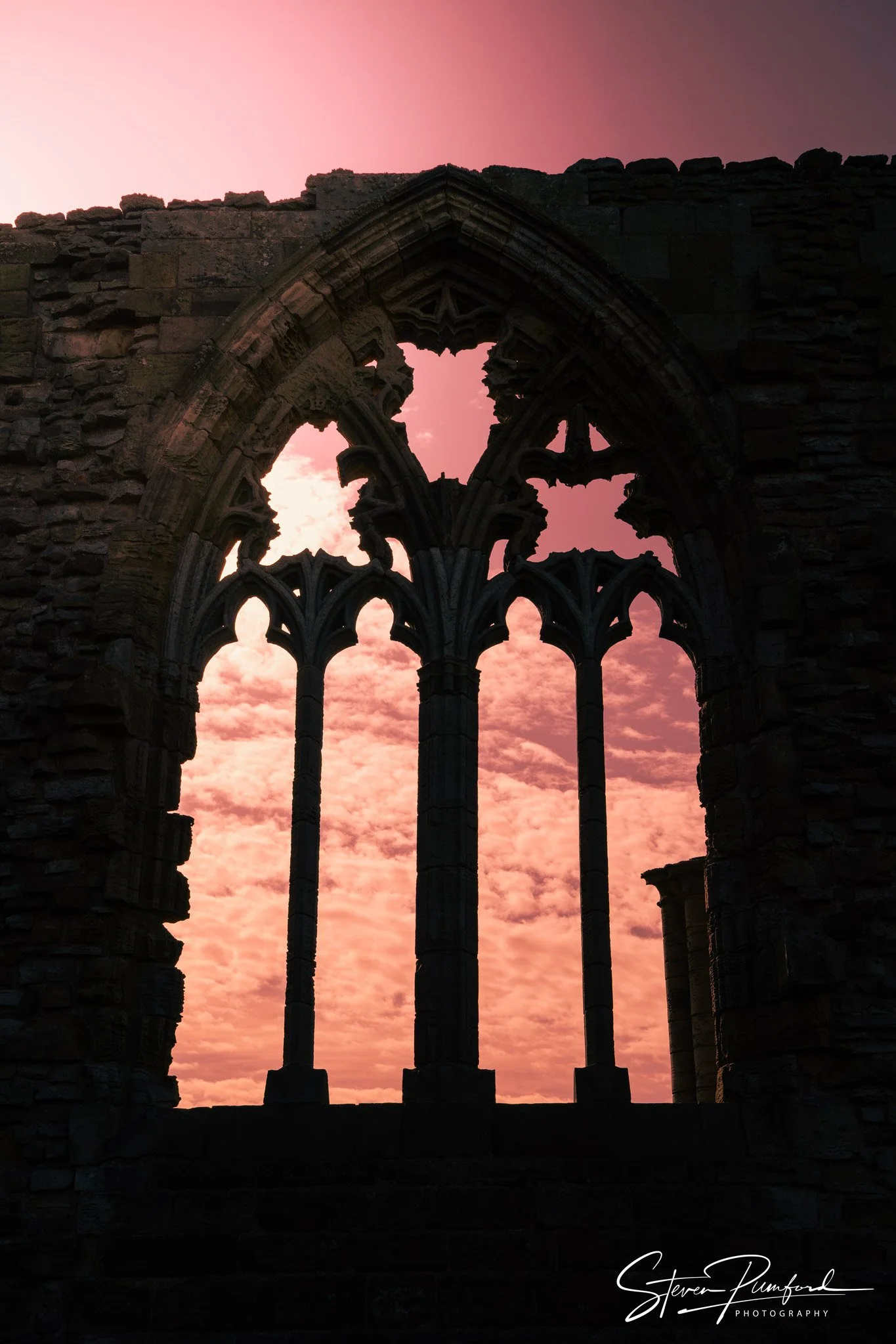 Silhouette of ancient stone window ruins with intricate gothic arches against a pink and orange sunset sky.