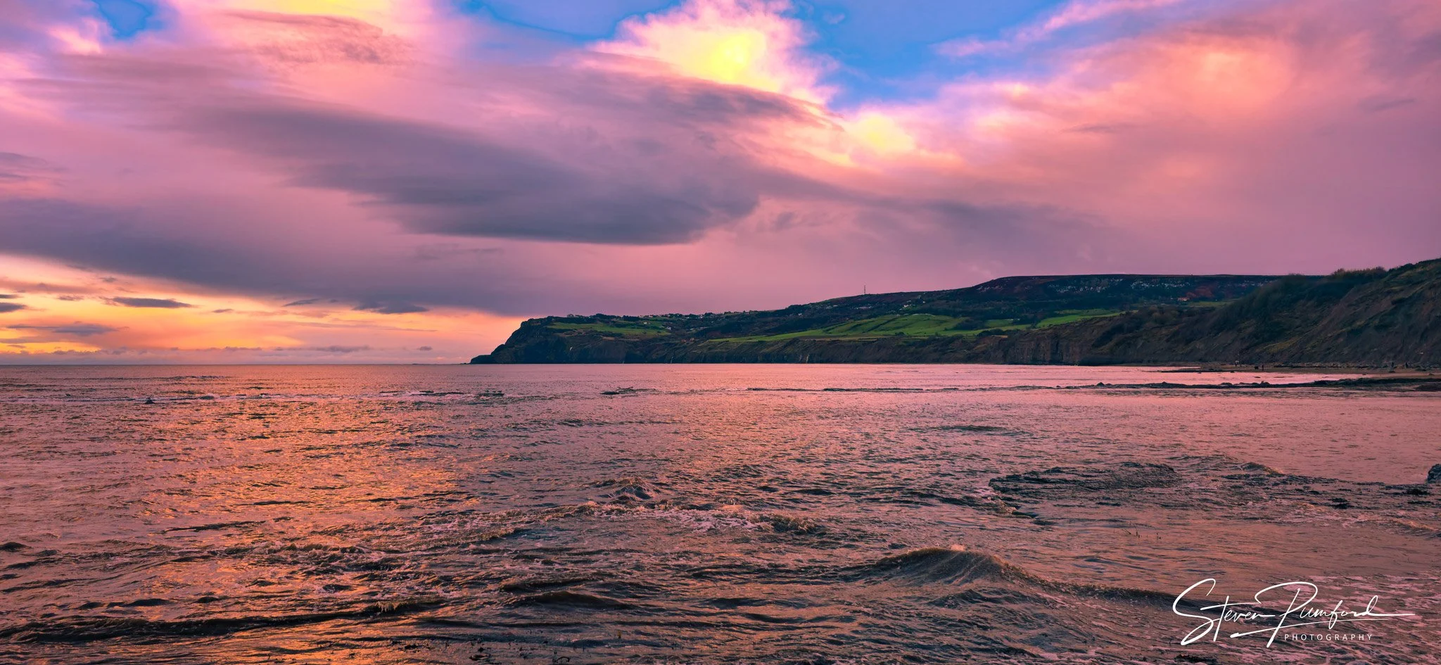 A beach at sunset with a colorful sky of pink, purple, and yellow hues, calm water, and green hills in the distance.