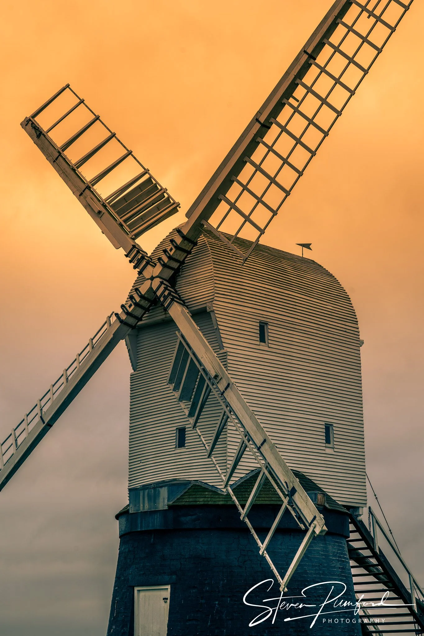 A traditional windmill with white wooden blades and a dark brick base, set against an orange sky at sunset or sunrise.