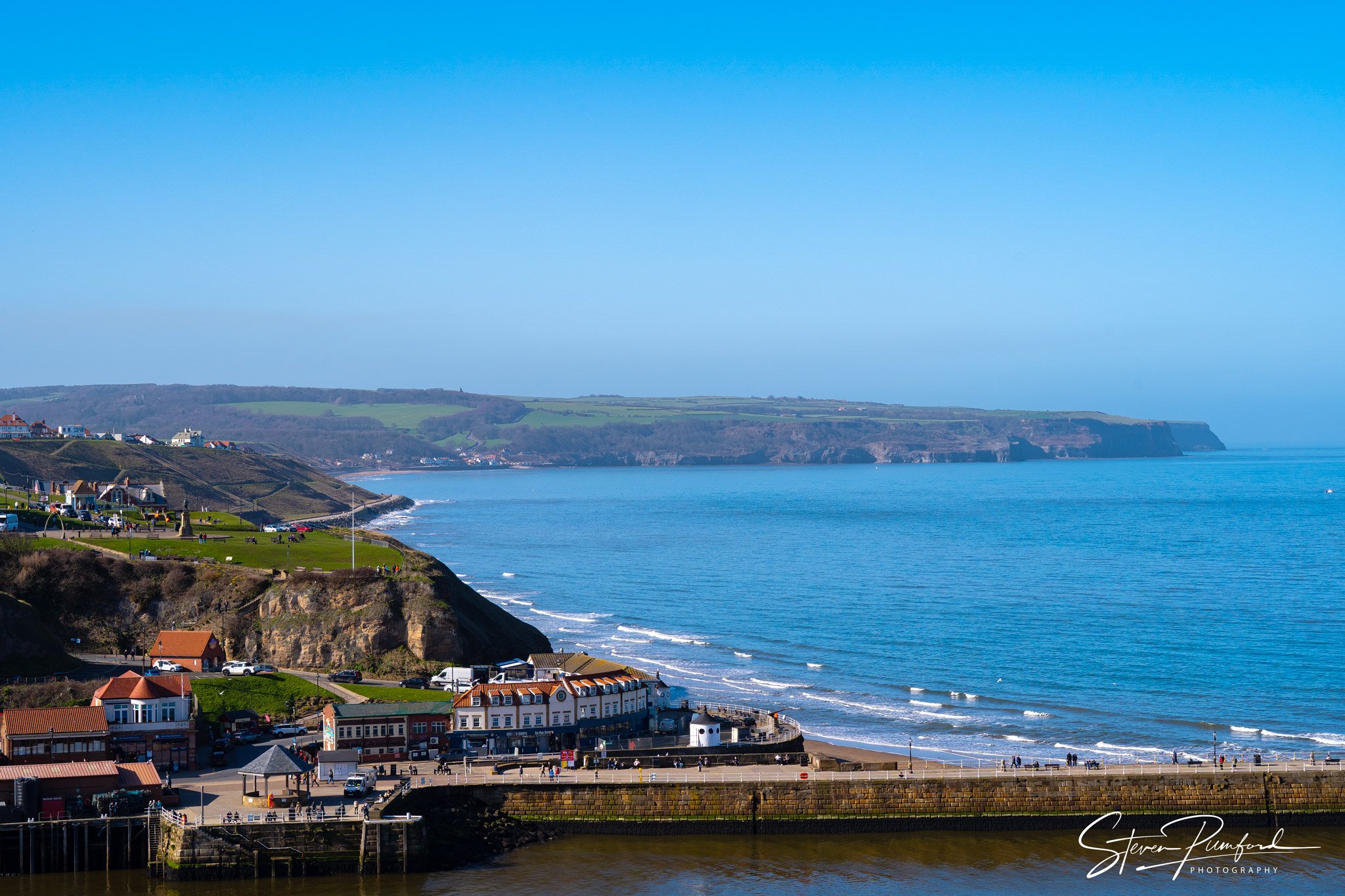 Coastal town with colorful houses along the shoreline, overlooking the ocean with cliffs in the background, under a clear blue sky.