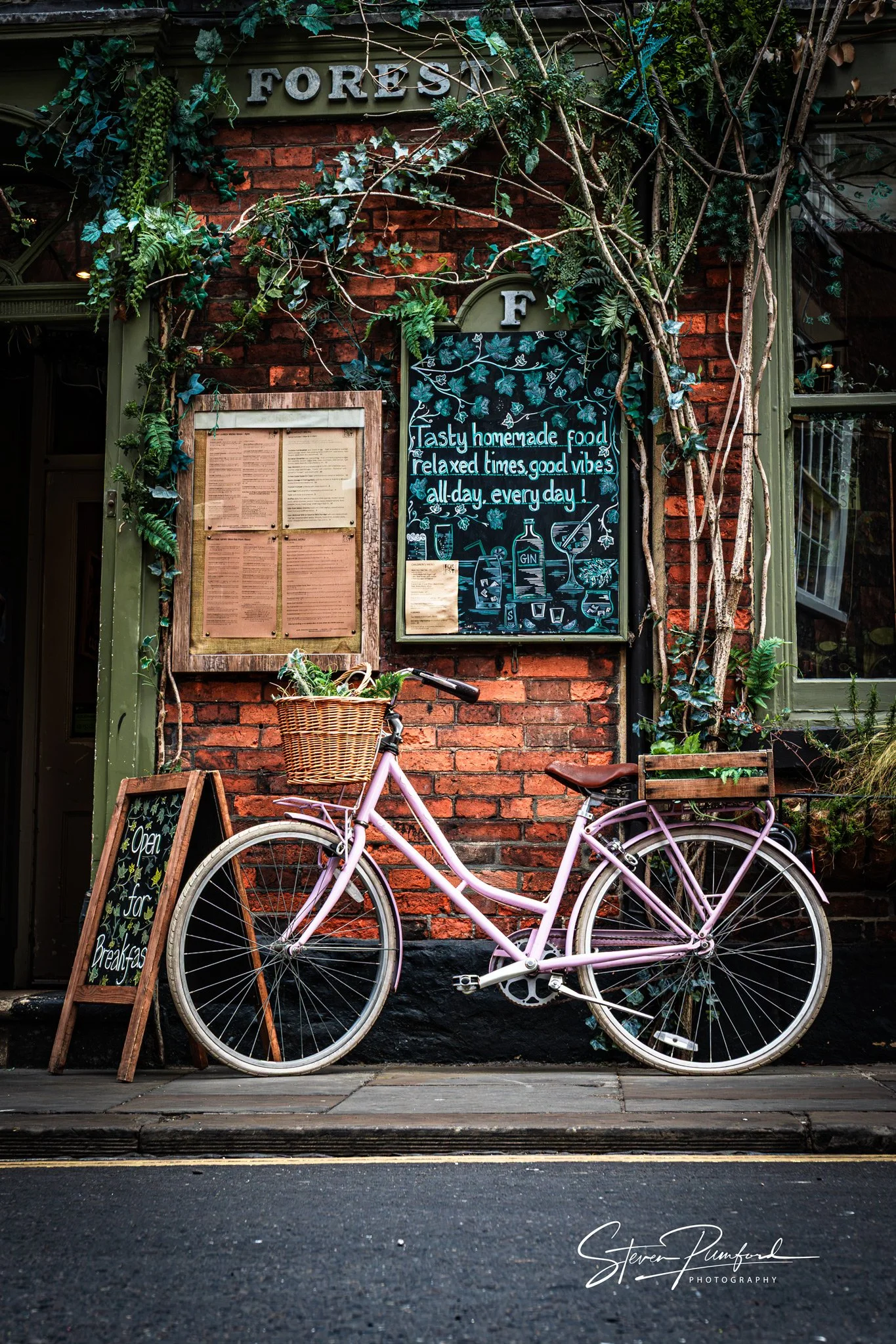 Pink bicycle with wicker basket parked in front of a brick wall decorated with green plants and signs, including a blackboard with recipes and a sign that says 'FOR' and 'Tasty homemade food relaxed times good vibes all-day every day!'