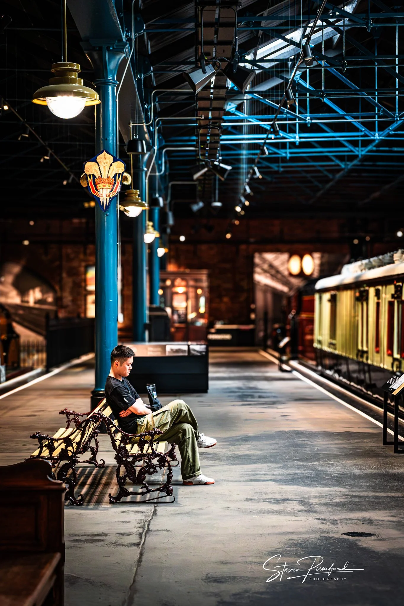 A person sitting on a bench in a train station, looking at their phone, with an urban interior and train on the platform in the background.