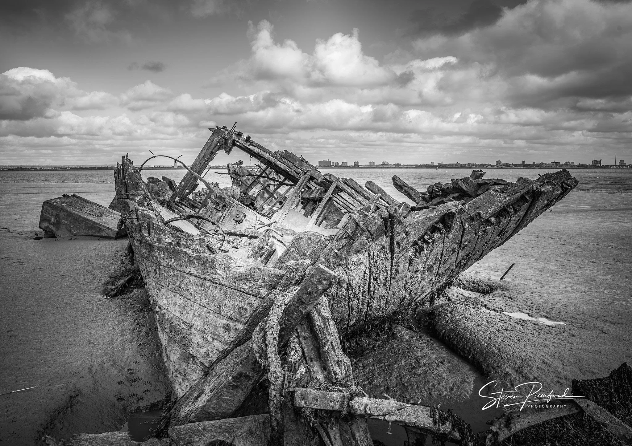 Black and white photo of a wrecked ship stranded on a sandy beach with a city skyline in the background and a cloudy sky overhead.