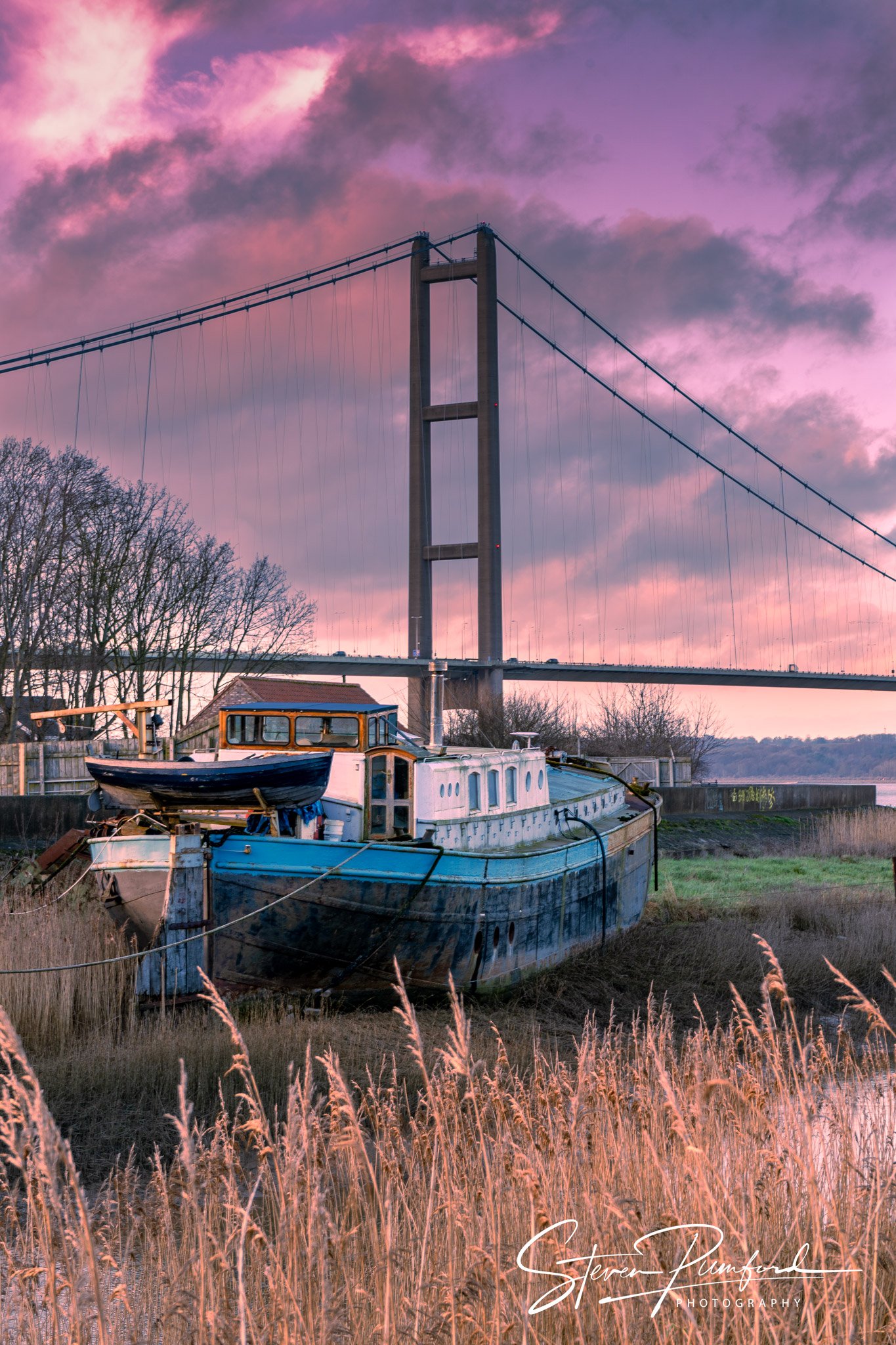 A vintage boat resting on the riverbank with tall grasses in the foreground and a bridge with suspension cables against a pink and purple sky at sunset.