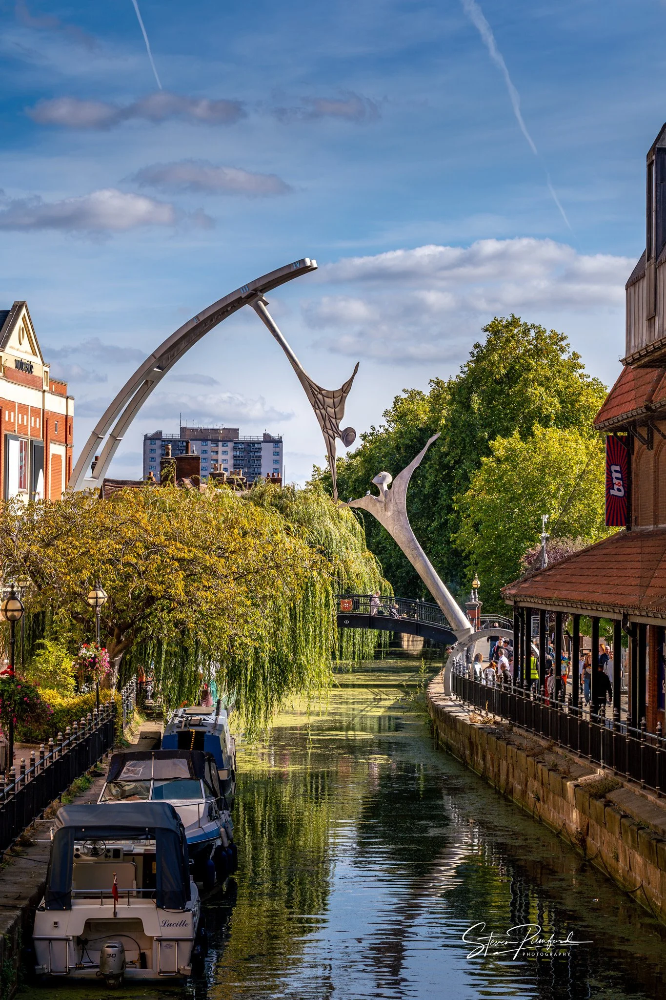 River Witham in Lincoln with boats docked along the side, surrounded by lush greenery and trees, with a bridge and buildings in the background on a sunny day with scattered clouds.