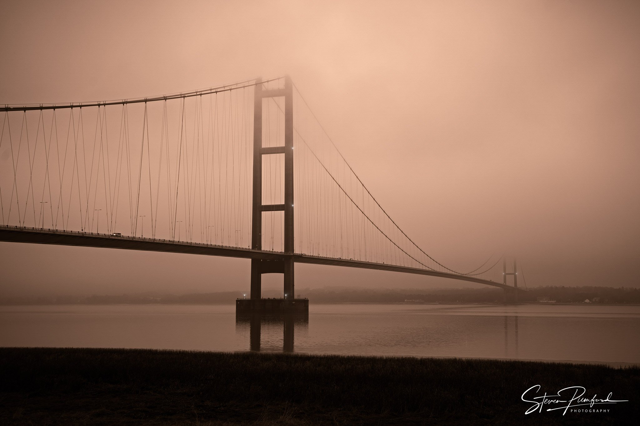 A foggy scene of a large suspension bridge over a body of water, with the bridge's towers barely visible through the mist.