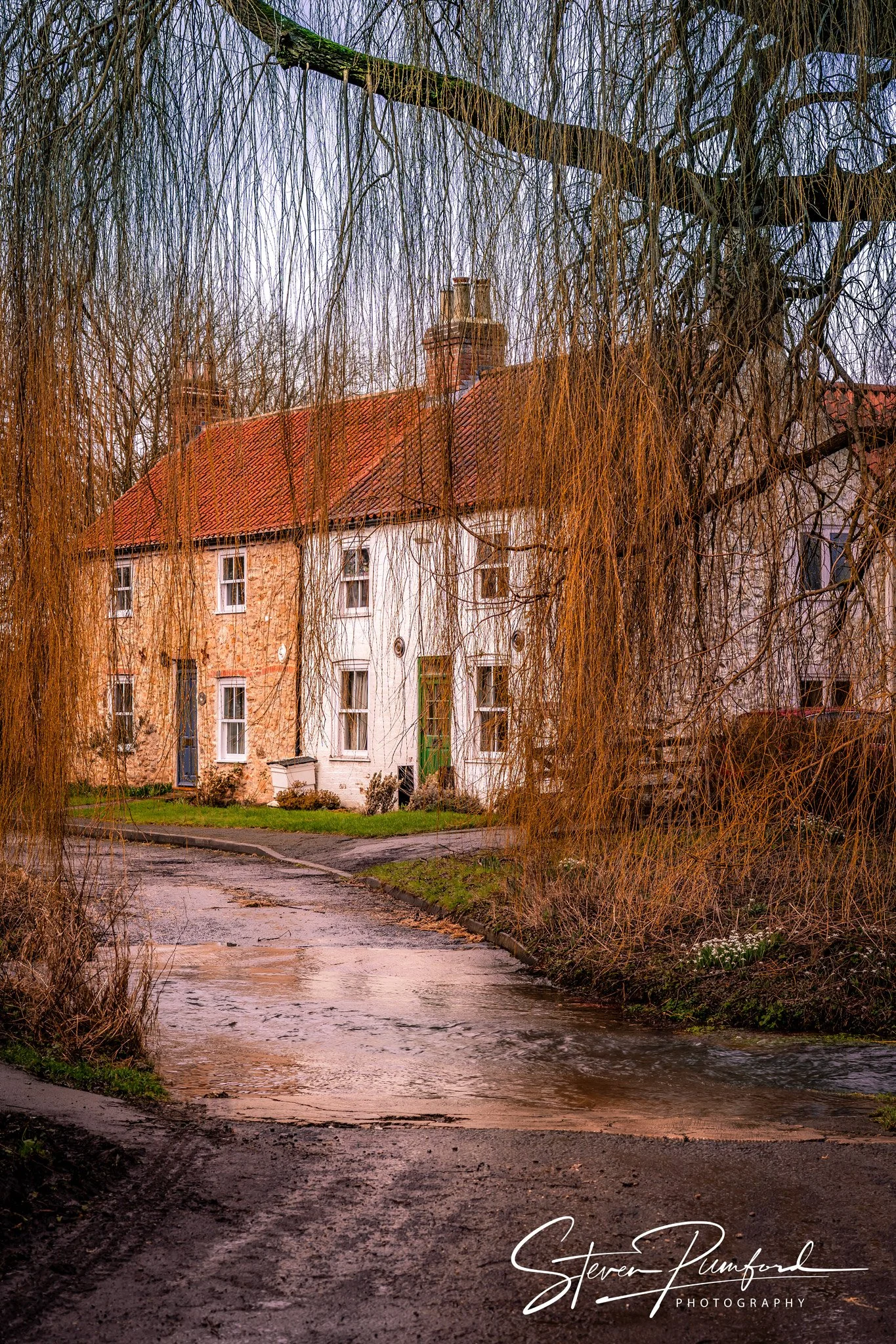 A scenic view of a house with a red tiled roof, surrounded by leafless trees, and a small creek in the foreground, with an overcast sky.