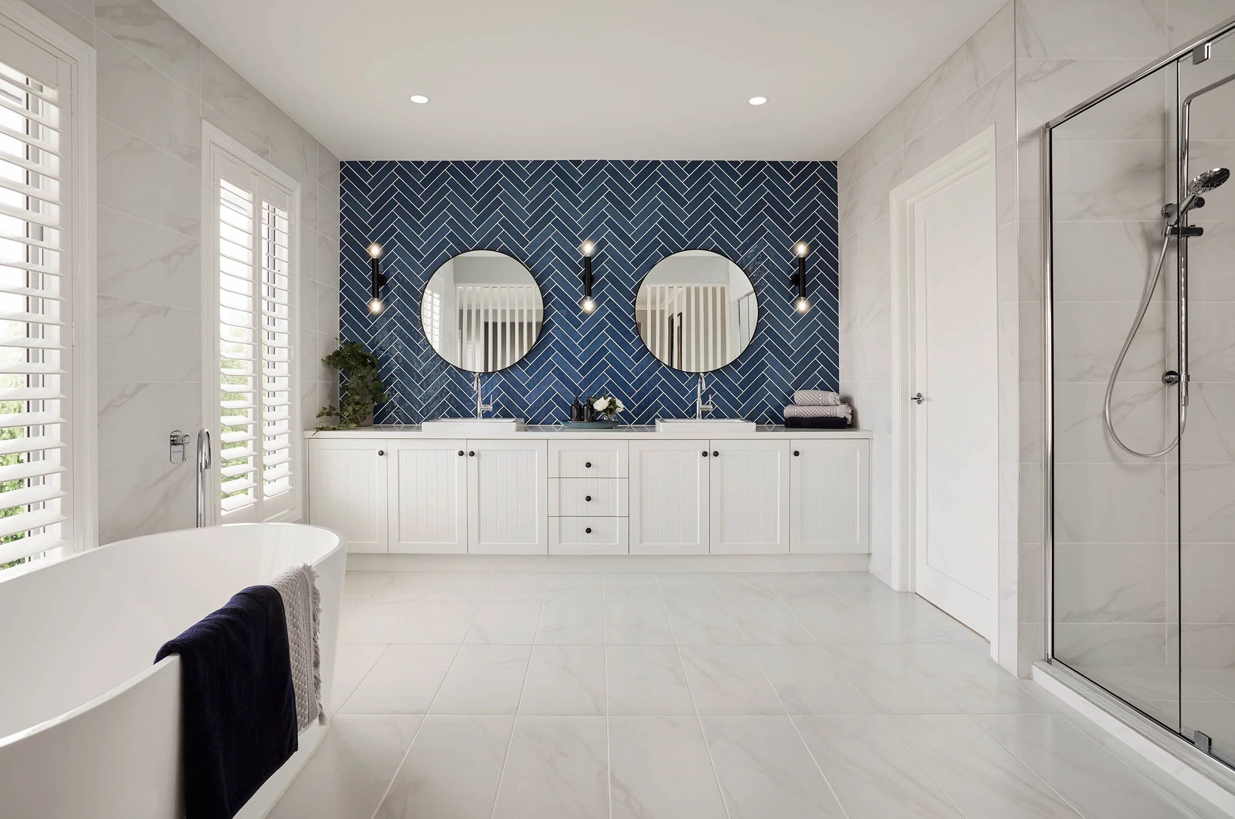Modern bathroom with white cabinets, two round mirrors on a blue tiled accent wall, and a walk-in shower with a glass door and chrome fixtures.