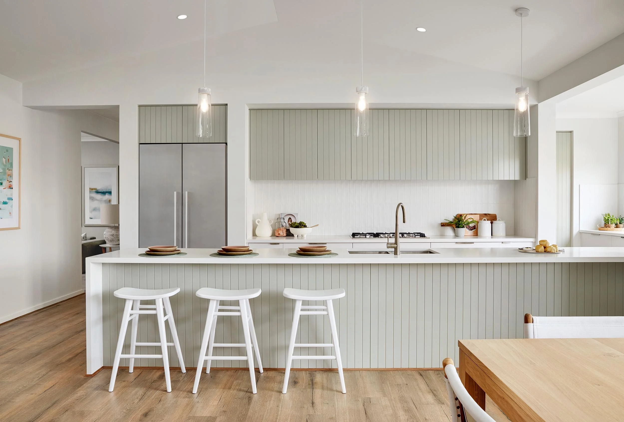 Modern kitchen with white island counter, three bar stools, light green upper cabinets, stainless steel refrigerator, and wooden dining table, decorated with potted plants and artwork.