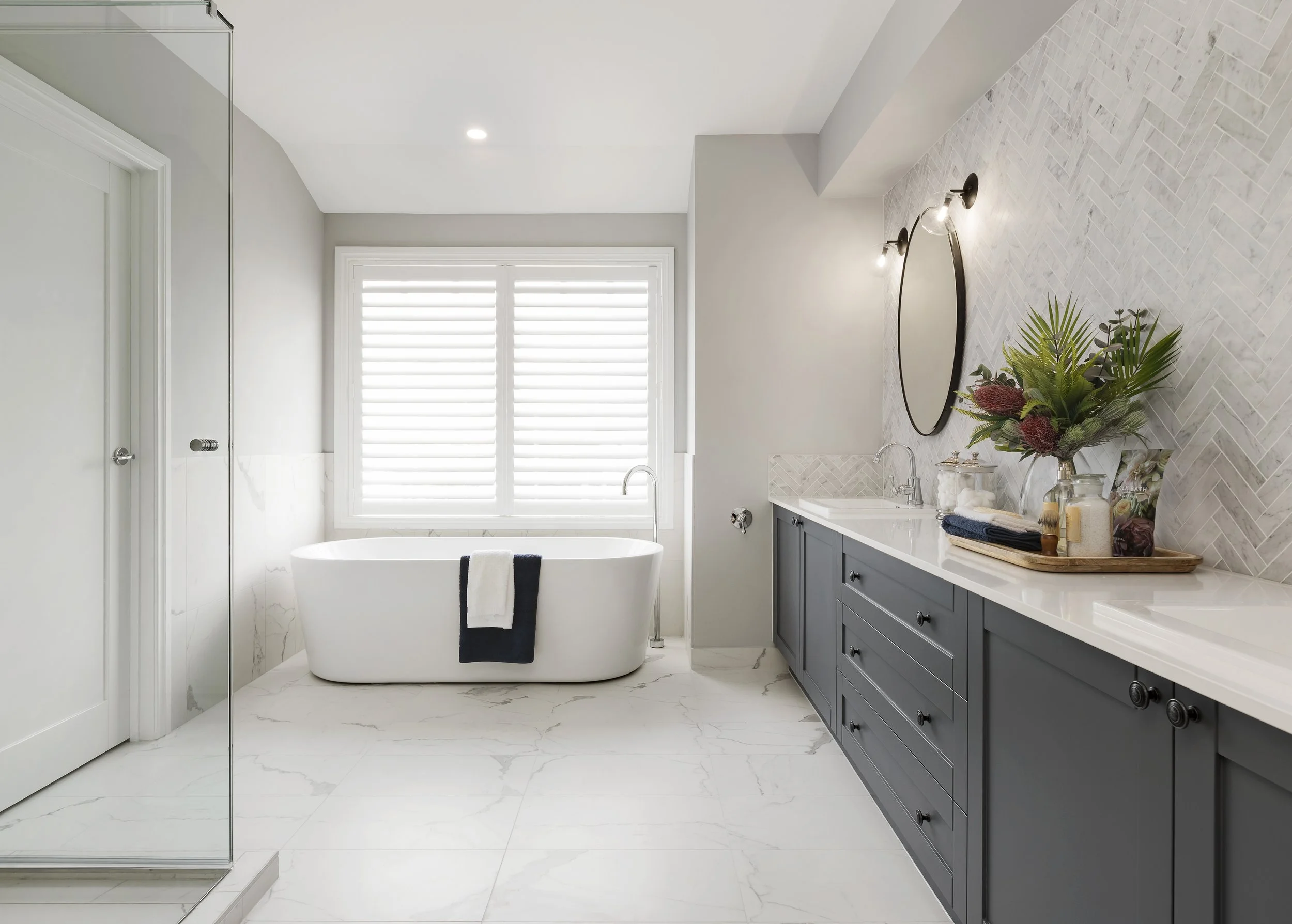A modern bathroom with a white freestanding bathtub in front of a window with white plantation shutters. To the right, there is a gray vanity with a white countertop, a round mirror, and a bouquet of green and purple flowers. The flooring and lower wall are made of white marble with subtle gray veining, and the upper wall has light-colored textured tile in a herringbone pattern. There are two black wall-mounted light fixtures above the mirror, and a glass shower enclosure is visible on the left.