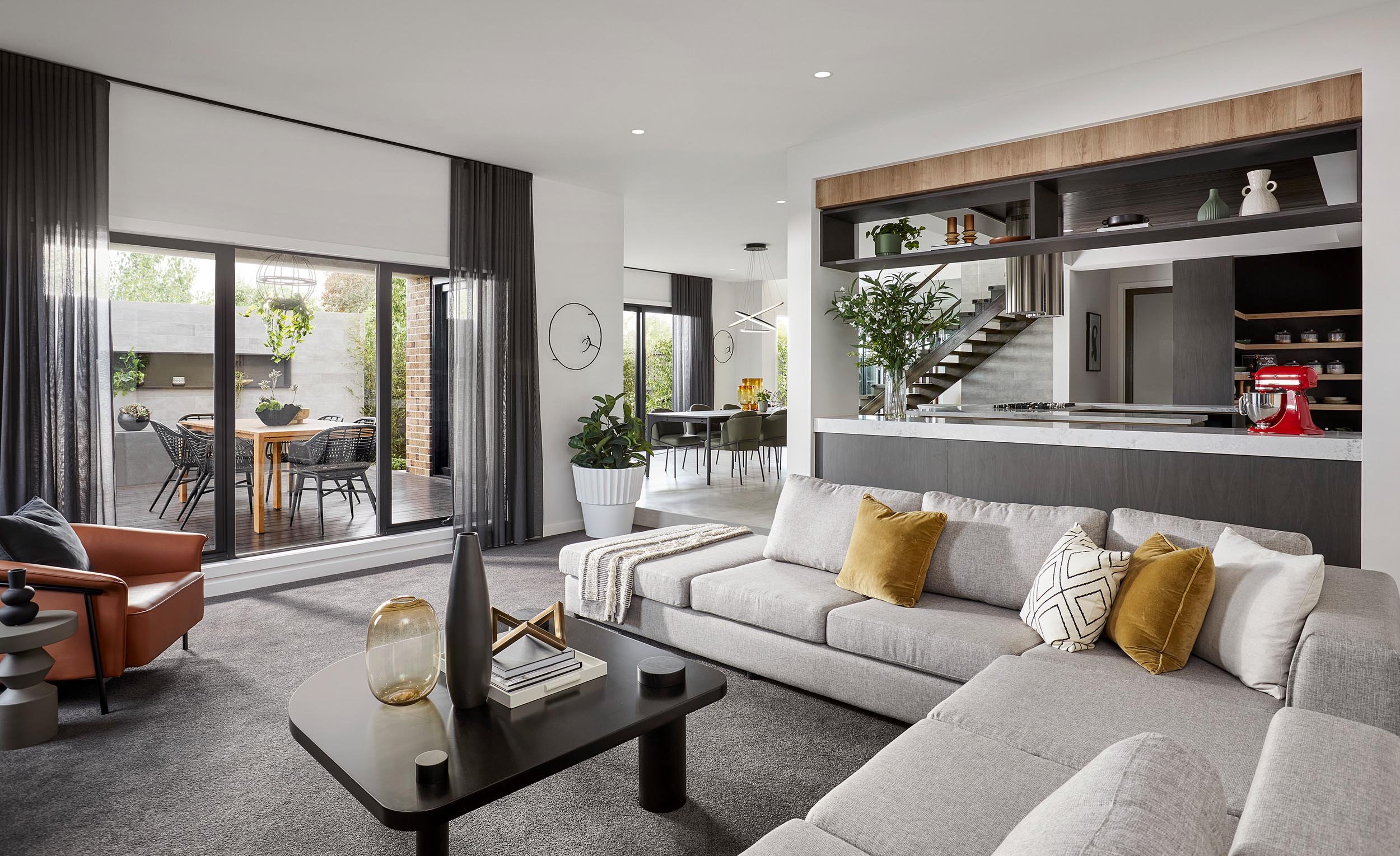 Modern living room with large window, tan sofa with pillows, black coffee table with decorative vases, adjacent dining area with greenery outside, open kitchen with staircase, pendant lighting, and decorative items on shelves.