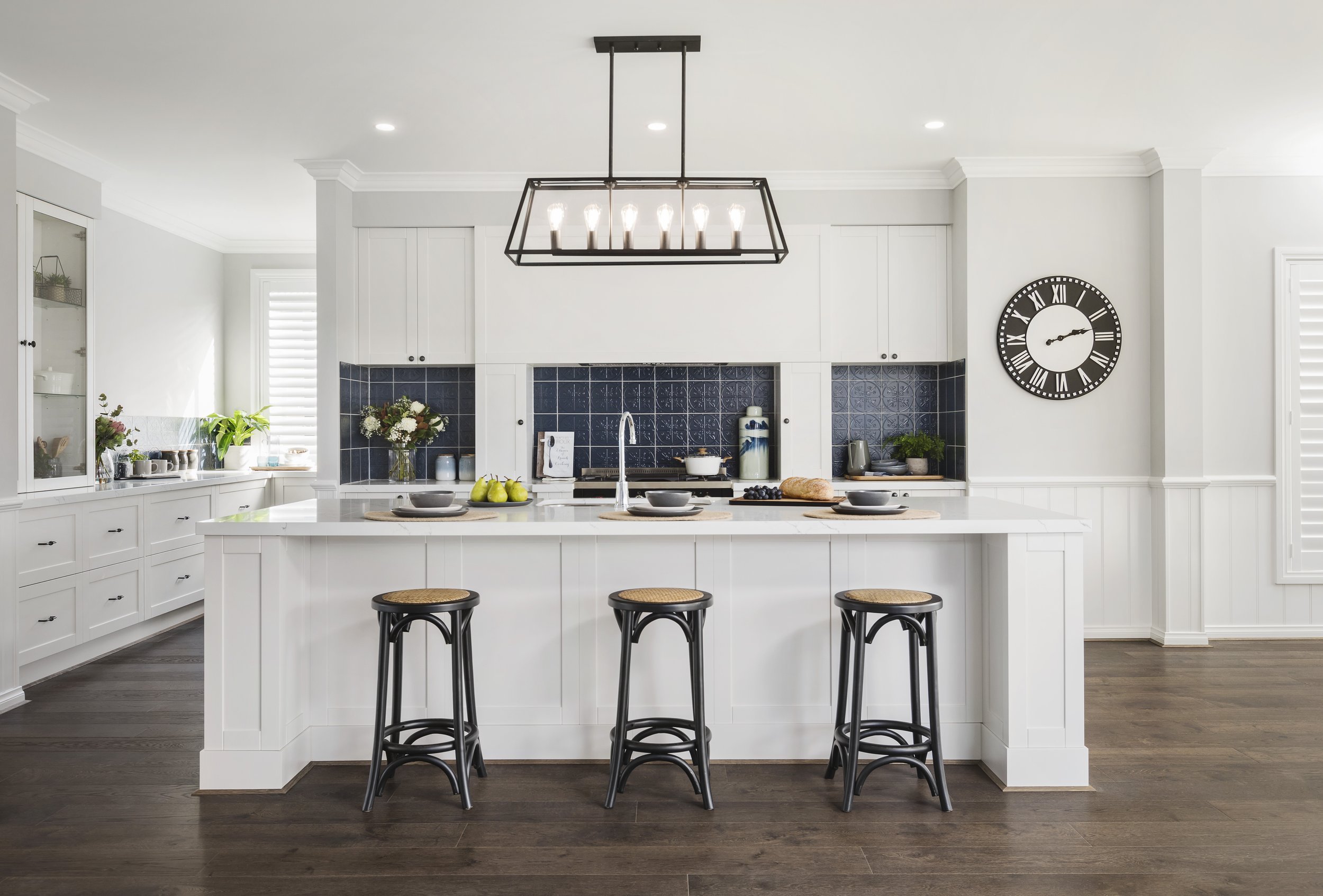 Modern white kitchen with island, black bar stools, dark wood floors, black and white clock, and black chandelier with exposed bulbs.