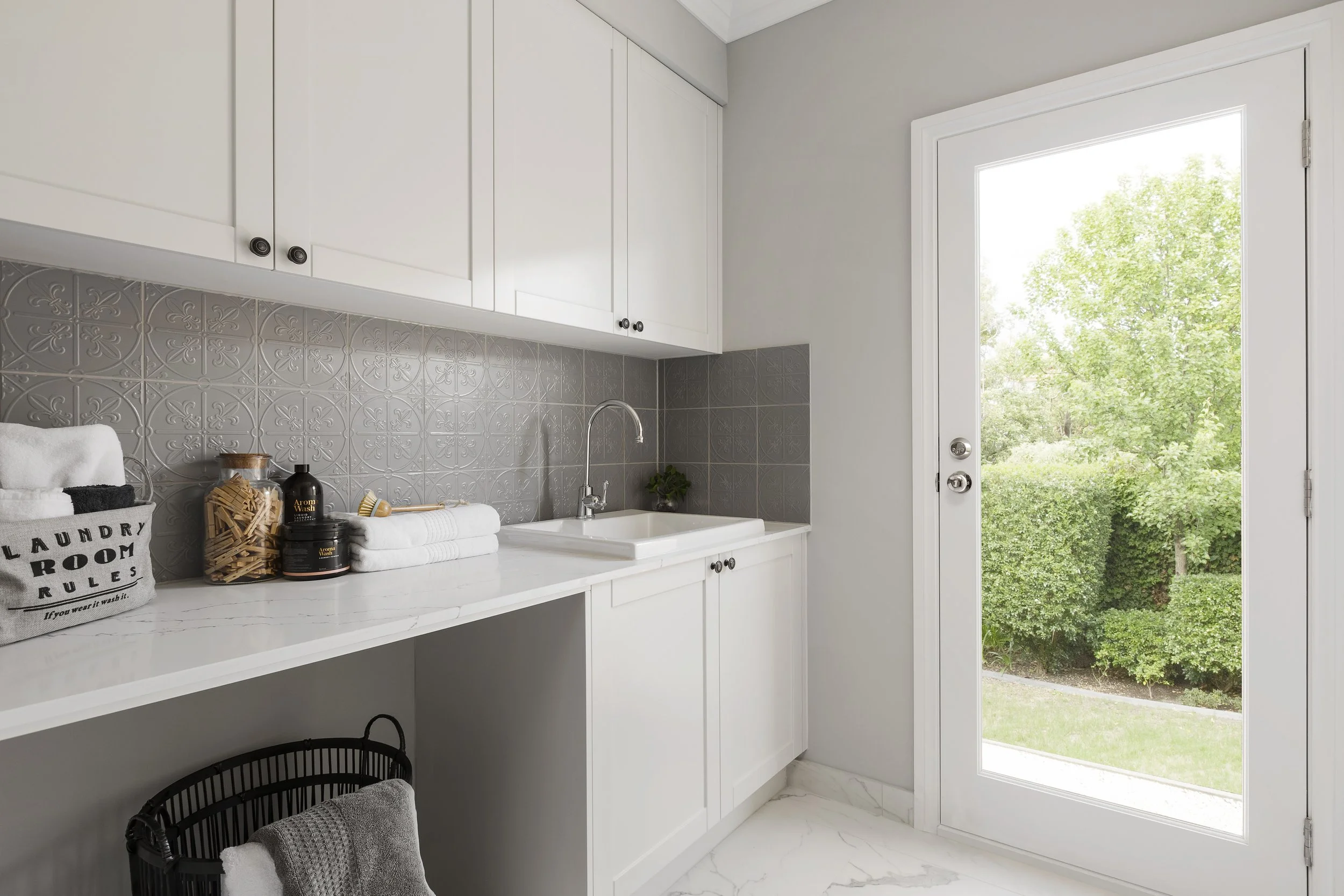 Kitchen with white cabinets, gray patterned backsplash, white countertop, and a door leading outside to a view of green trees.