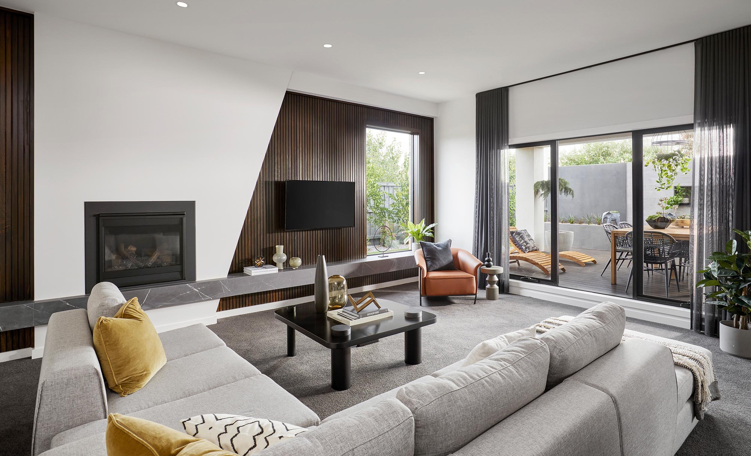 Modern living room with a fireplace, flat-screen TV, beige sectional sofa with yellow pillows, coffee table with decor, and sliding glass door leading to a balcony with outdoor furniture and greenery.