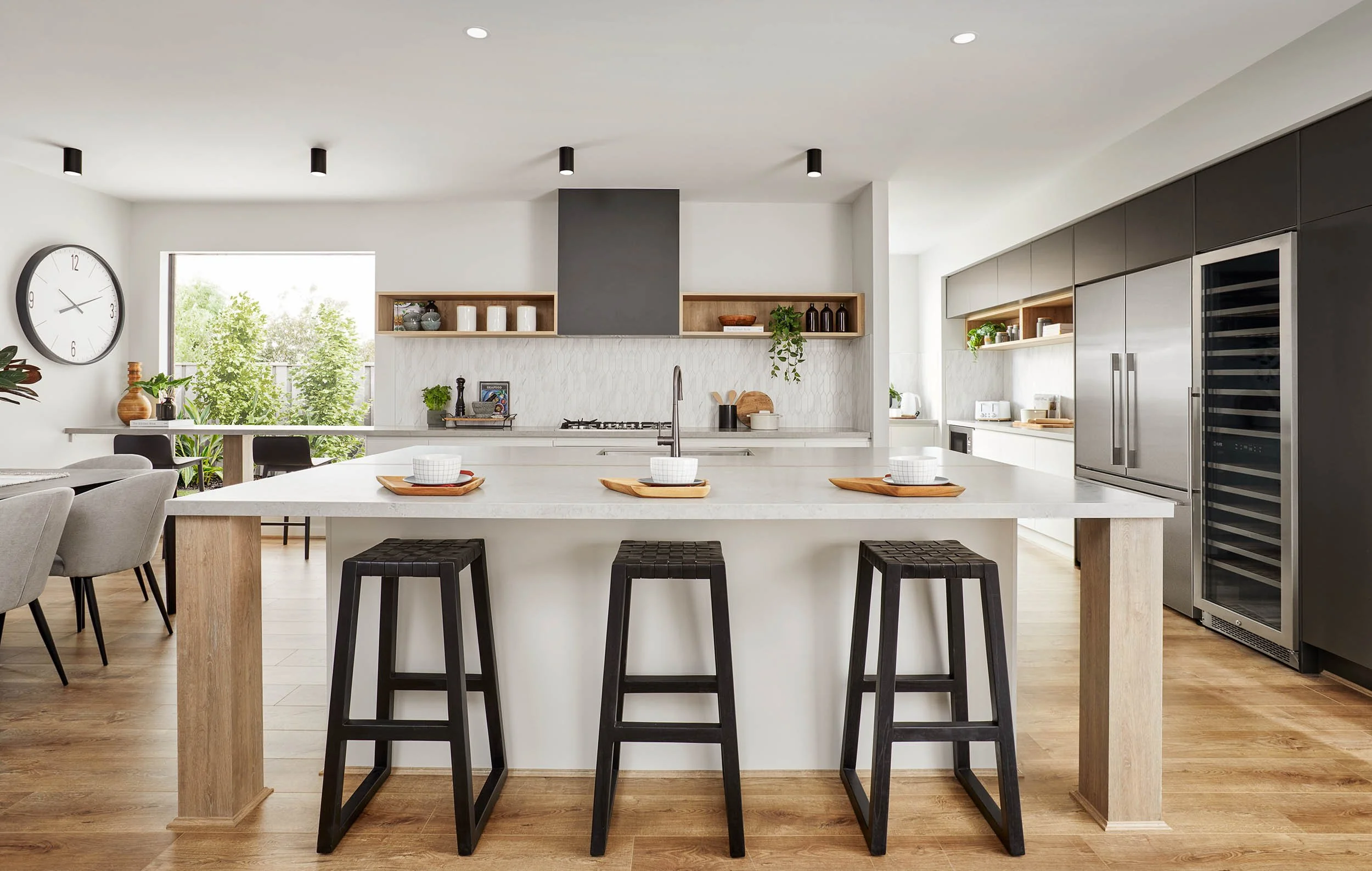 Modern kitchen with white island, black stools, open shelves, stainless steel appliances, and large window with greenery outside.