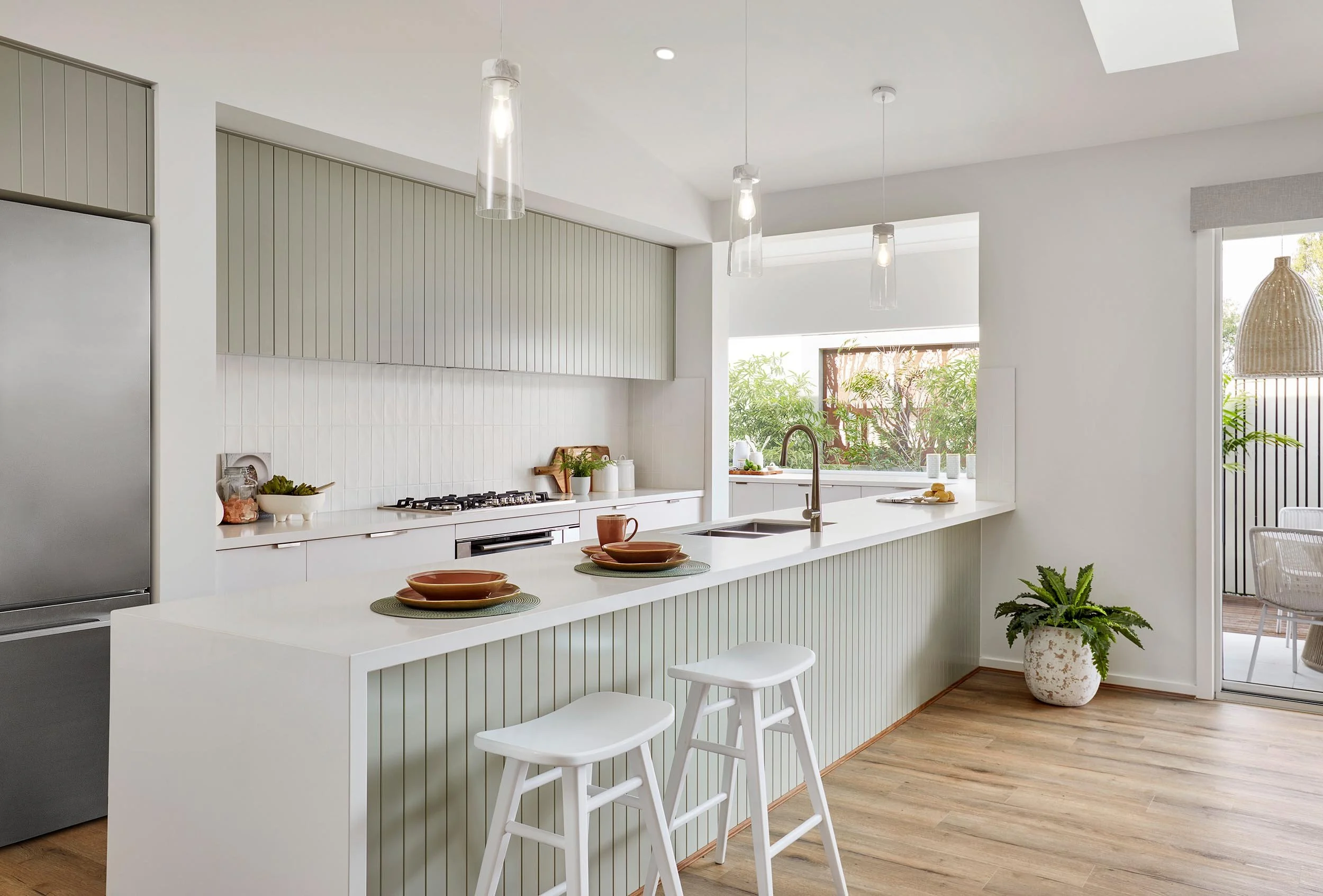 Modern white kitchen with barstools, pendant lights, and large window with greenery outside.