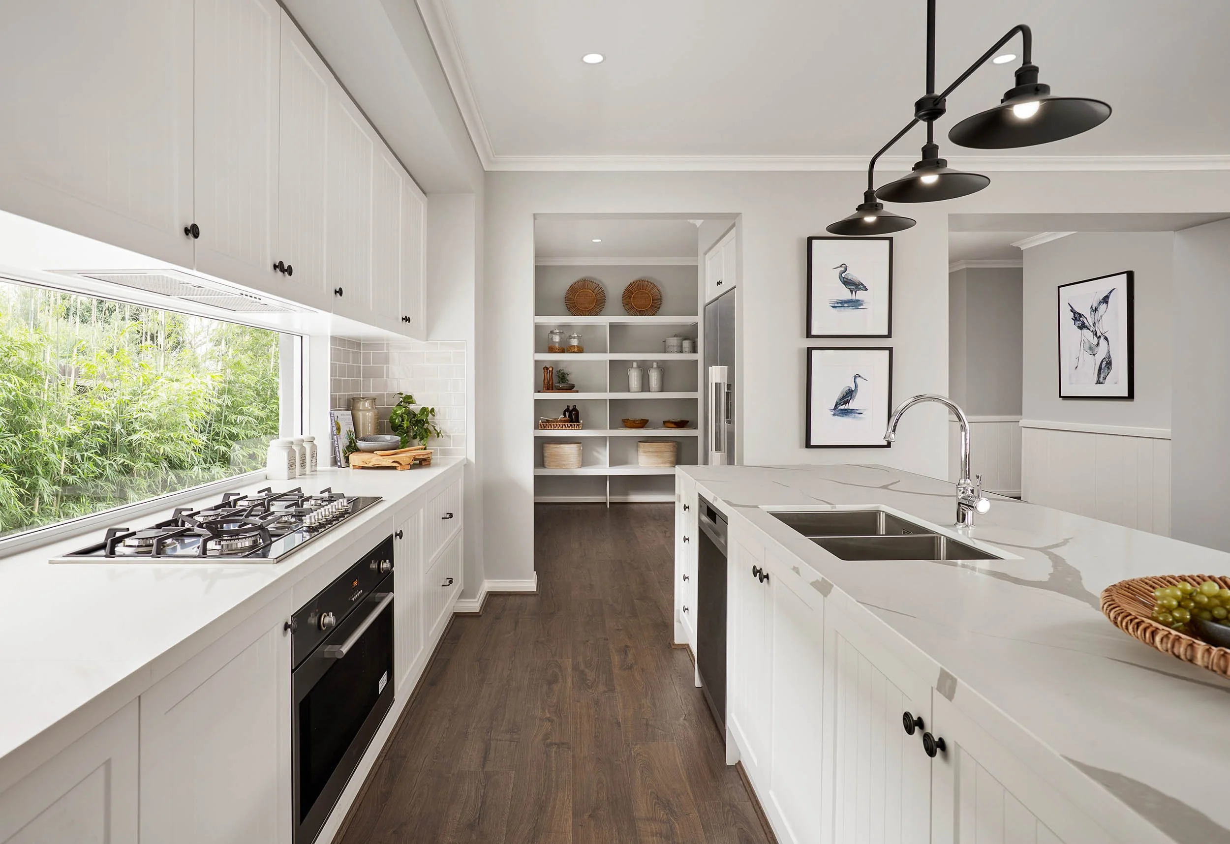 Modern white kitchen with island, black pendant lights, and framed bird artwork, large window with green foliage outside.