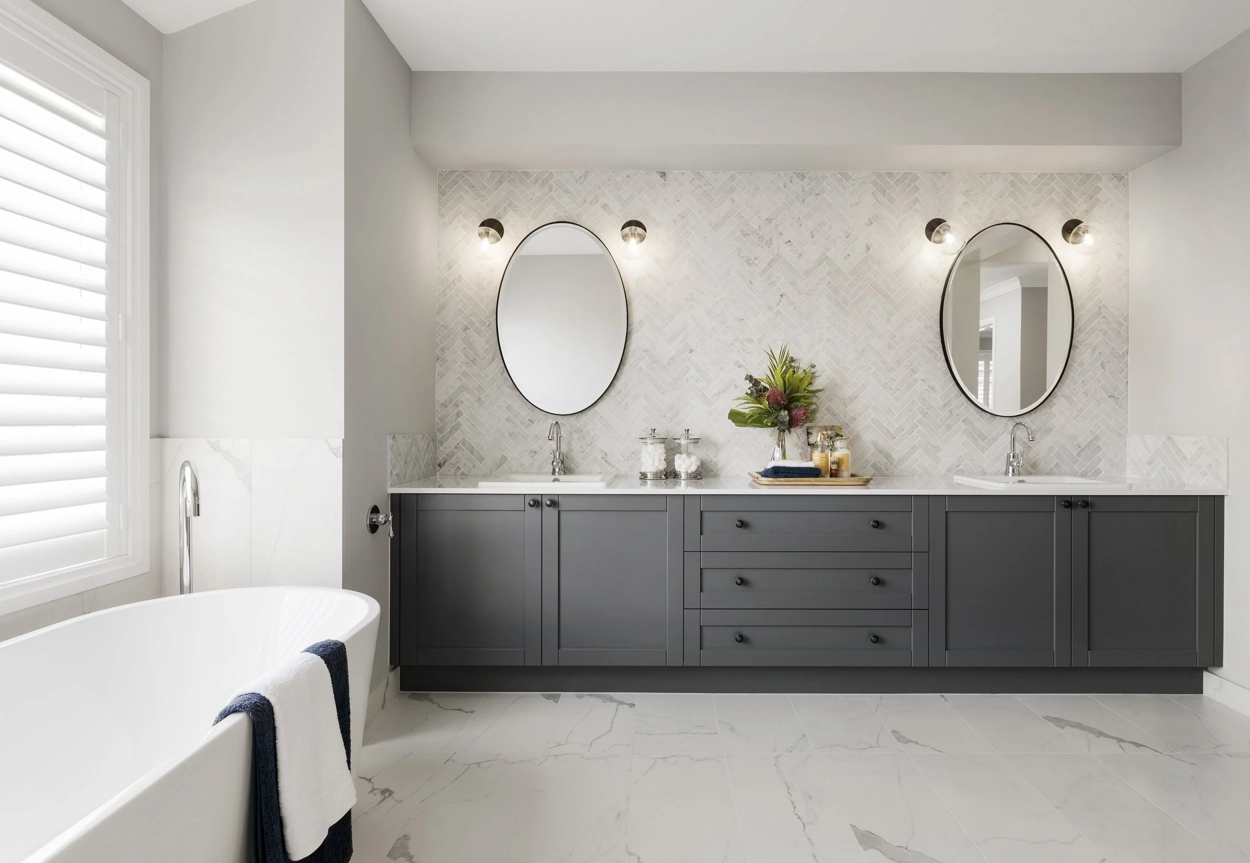 A modern bathroom with a double vanity with grey cabinets, two oval mirrors with black frames, and wall-mounted lights. The vanity has a decorative tray with plants and toiletries. There is a bathtub with a towel hanging on the edge and a window with